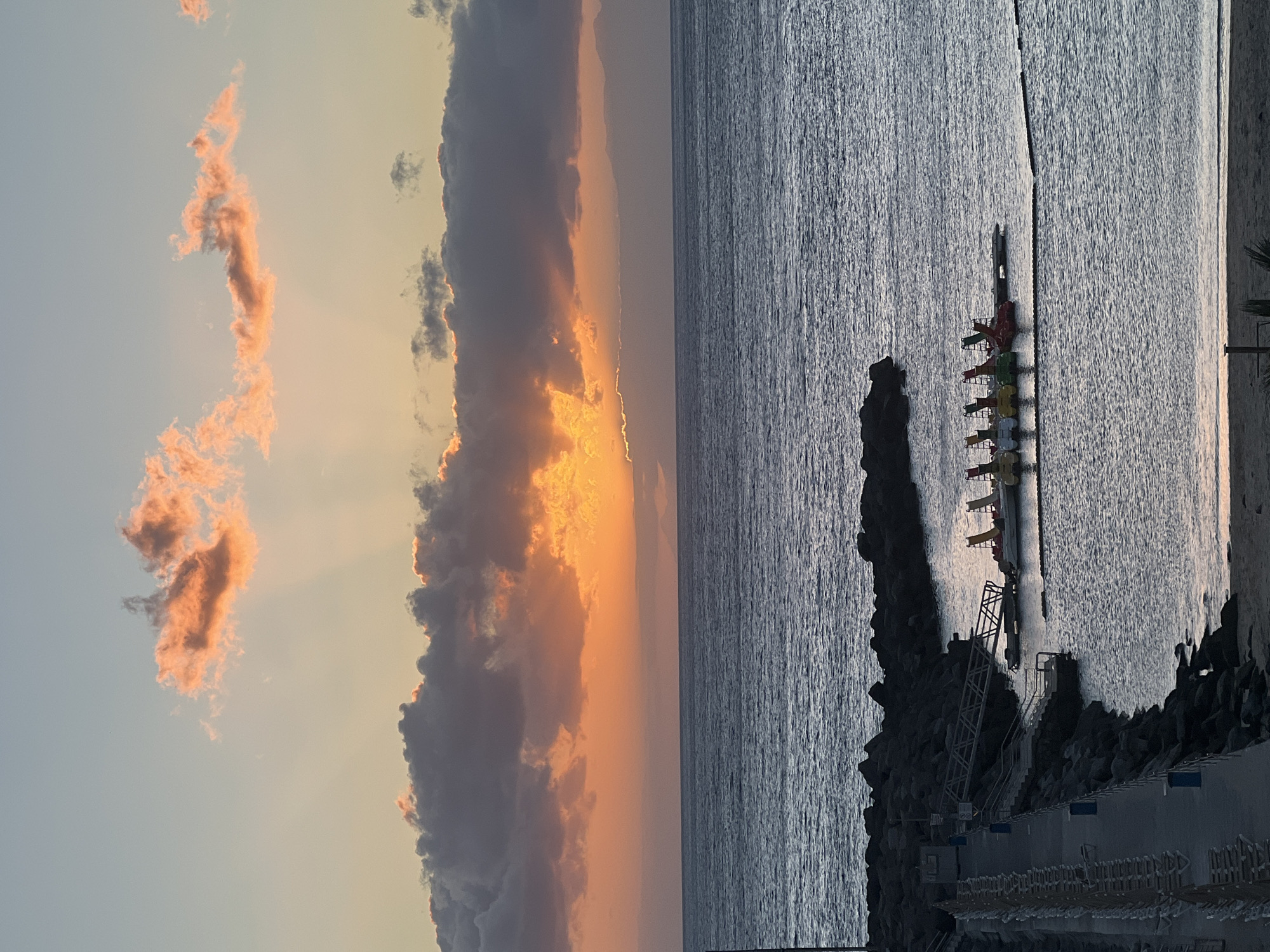 A sideways shot shows a sunset over the ocean in Mogán, Spain. On the left, pink-tinged clouds are illuminated by the setting sun, partly obscured by a dark, heavy cloud formation. The sun's rays break through these clouds. The right half of the image features the ocean's surface, rippled by the wind. A pier made of large rocks extends into the water, with a staircase leading down to a concrete walkway marked with blue rectangles. A row of brightly colored paddle boats is docked along the pier. A palm tree is barely visible on the bottom right corner.