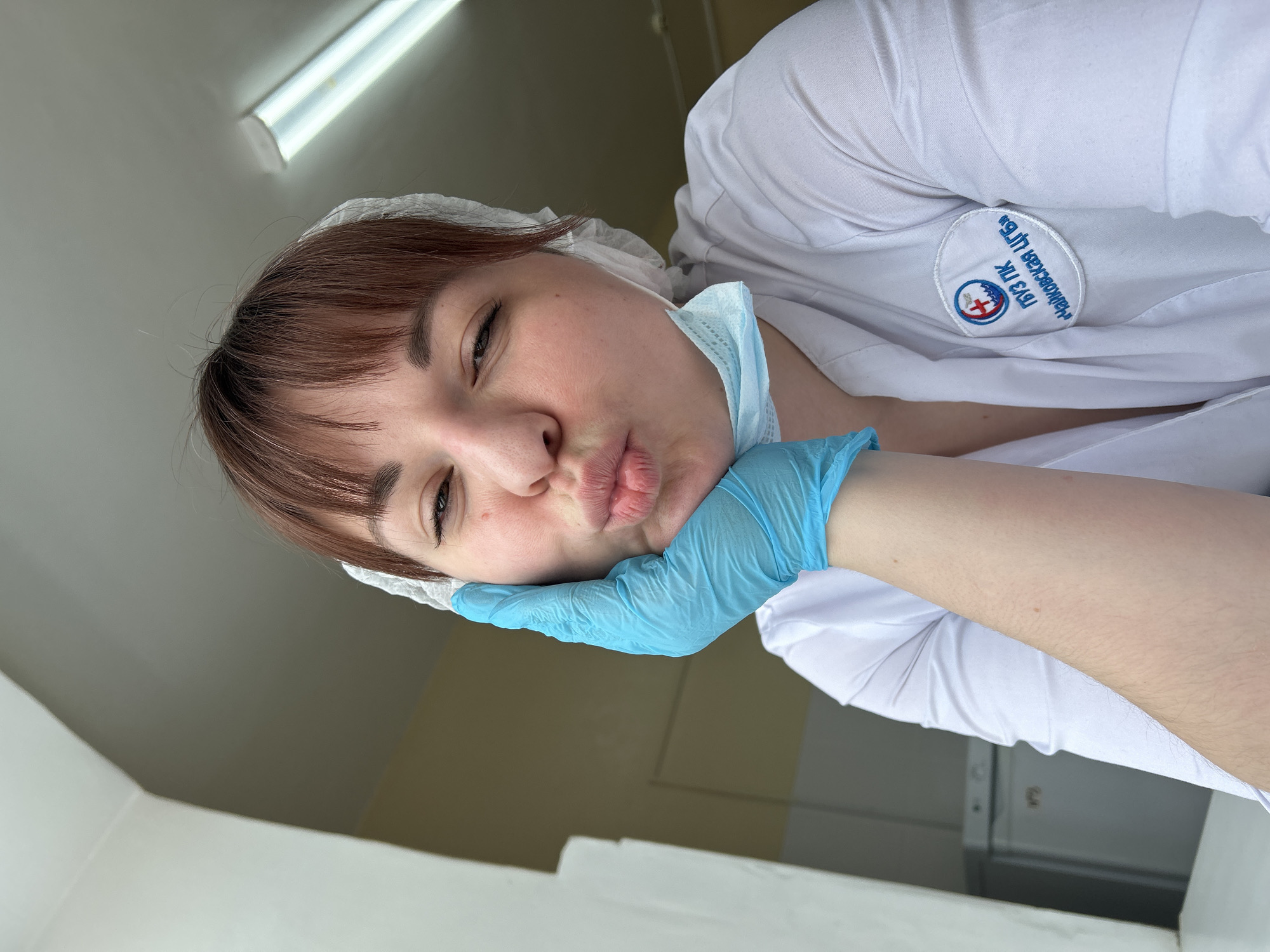 The scene depicts a female medical professional indoors at a facility in the village of Foky, Chaykovsky District, Perm Krai, Russia. She is shown from the upper torso upward, with reddish-brown hair and bangs, partially covered by a white disposable hairnet. She is wearing a white medical coat or scrub top, bearing an embroidered patch on the left chest identifying the "Tchaikovsky Central City Hospital" (ГБУЗ ПК "Чайковская ЦГБ"). A light blue disposable surgical mask is hanging below her chin, and her left hand is covered by a light blue disposable nitrile glove, resting against her left cheek. She is looking directly at the camera with a playful expression, featuring puckered lips and partially closed eyes. The background consists of light-colored walls, a ceiling-mounted fluorescent light fixture in the upper right, and a glimpse of a metallic appliance, possibly a refrigerator, on the left.