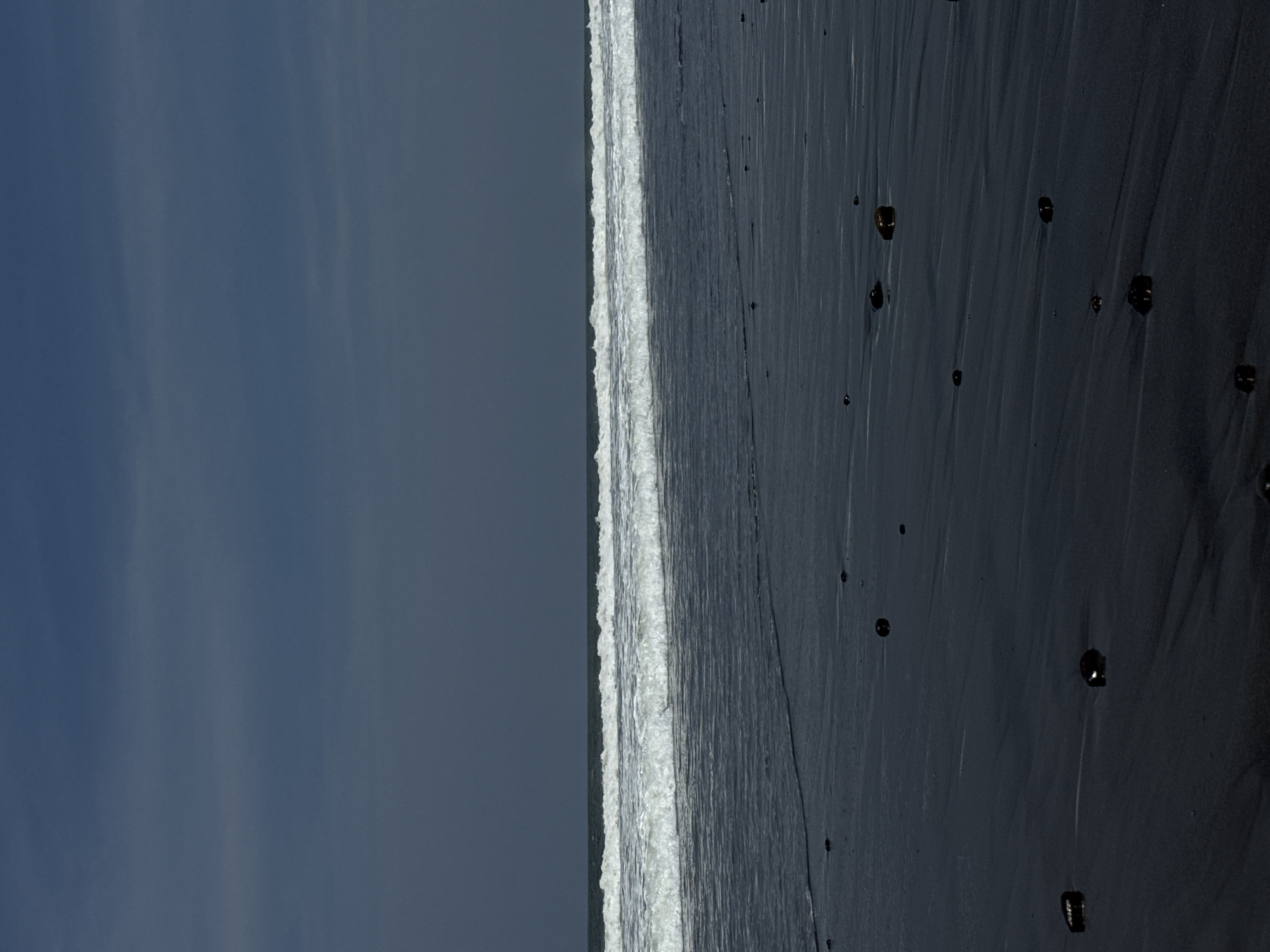 A vertical image depicts a dark sandy beach meeting the ocean under an overcast sky in Sudimara, Indonesia. The upper half of the frame shows a dark blue-grey sky with subtle cloud variations. Horizontally across the mid-ground, a line of white, breaking ocean waves extends, indicating an active tide. The water immediately in front of the waves appears dark and reflective. The lower half of the image consists of the wet, dark, possibly black sand beach, exhibiting minor ripples and surface patterns from receding water. Scattered across the foreground and mid-ground of the beach are numerous small, dark, rounded objects, consistent with pebbles or small rocks. No individuals, animals, or structures are visible.
