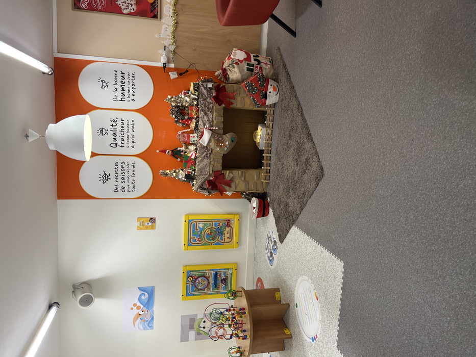 The image shows a brightly colored children's play area. A faux fireplace is decorated with Christmas decorations, including stockings and wrapped gifts. The walls are adorned with colorful graphics and signage in French, promoting fresh, seasonal food. Toys such as bead mazes and wall-mounted activity boards suggest a focus on early childhood education and entertainment. The flooring is a mix of gray carpet and interlocking foam tiles, offering a comfortable and safe environment for kids to play.