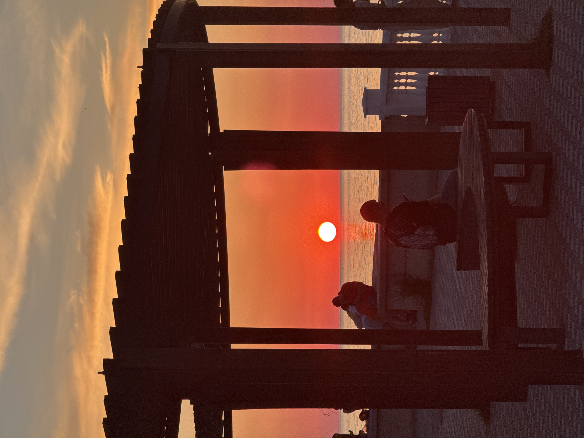In a city park, a serene sunset paints the sky with vibrant hues of orange and yellow. The sun, a luminous orb, casts a warm glow over the calm sea. A couple sits closely, silhouetted against the breathtaking backdrop, embracing the tranquility of the moment. In the foreground, a person sits on a bench, engrossed in their phone, while another stands nearby, observing the picturesque scene. The park, with its paved walkways and wooden benches, offers a peaceful respite from the urban hustle, creating a harmonious blend of nature and city life.