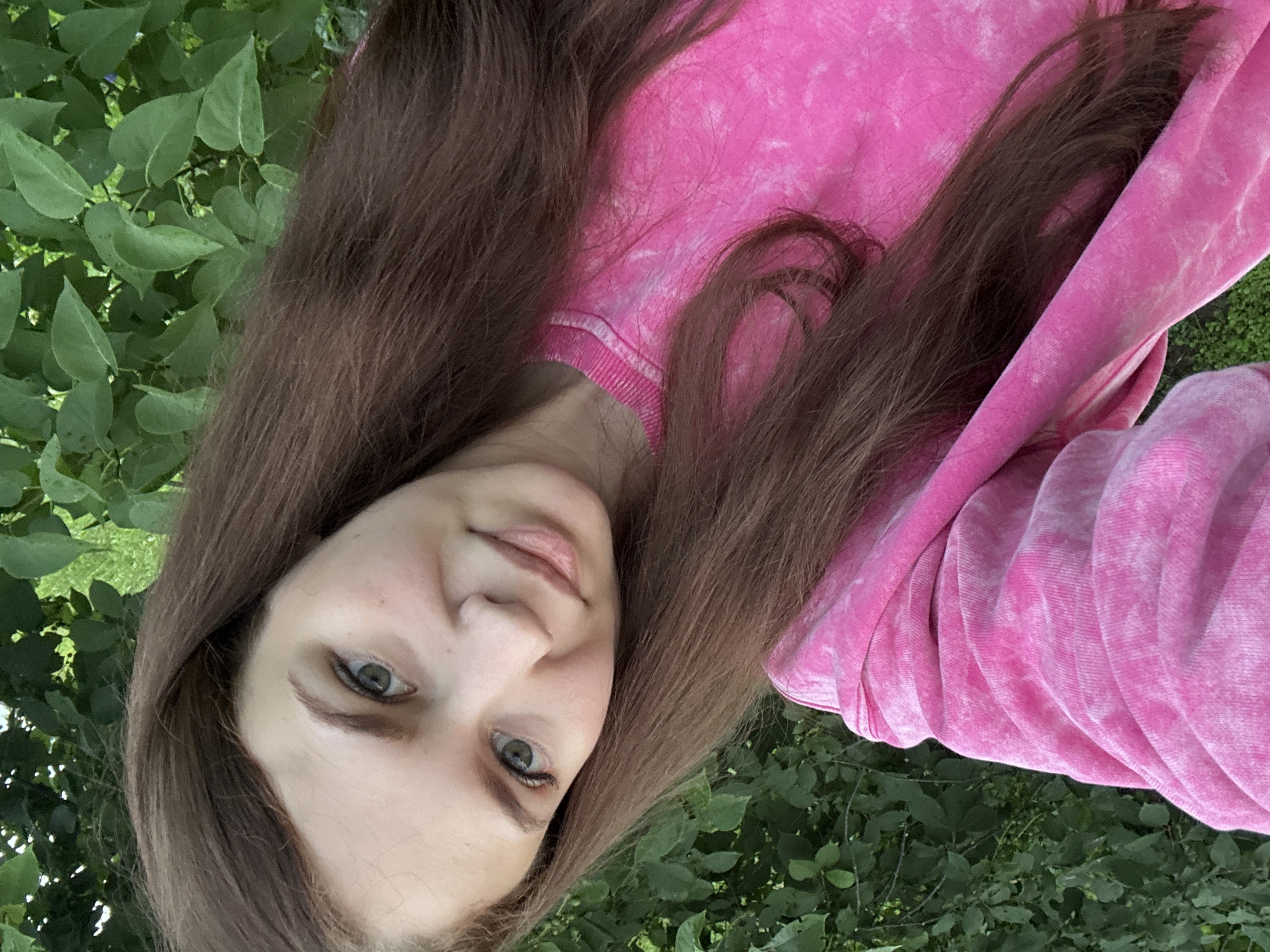 A young woman with long, brown hair and blue eyes is pictured outdoors, with her head tilted towards the camera. She is wearing a bright pink sweatshirt. The background consists of lush green foliage, suggesting a natural, possibly wooded or garden setting. The image is taken during daylight hours, as indicated by the bright lighting. The woman's expression appears relaxed and content. There is no visible text in the image.