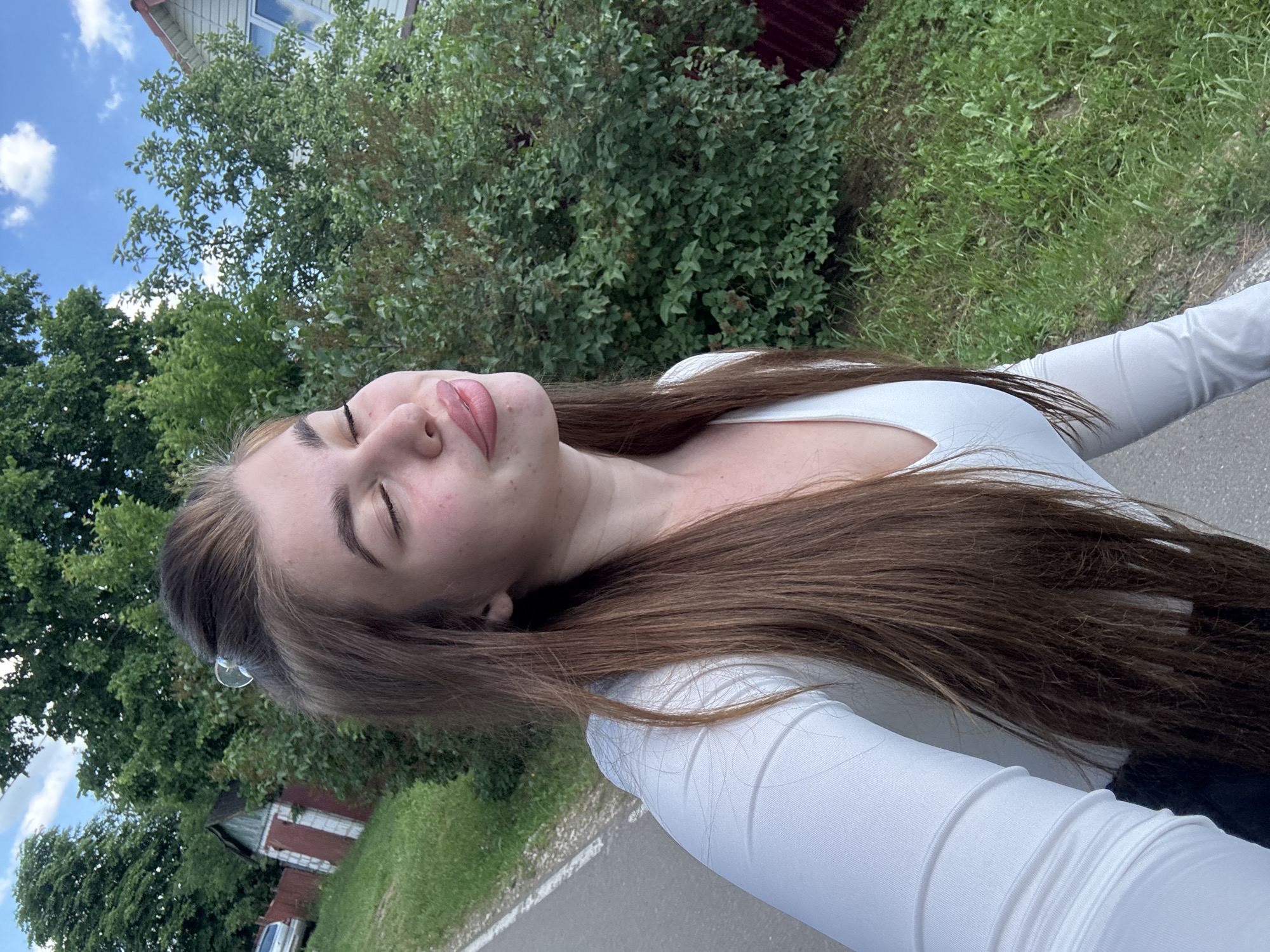 A young woman with long brown hair is taking a selfie outdoors in a rural setting. She is wearing a white long-sleeved shirt and appears to be smiling with her eyes closed. The background consists of lush green trees, grass, a gravel road with a white line, a small building with a red roof and white trim, and a fence. The sky is blue with scattered white clouds. The scene suggests a sunny day, possibly in a village or countryside near Мещёры, Russia, given the architectural style of the building and the natural landscape.