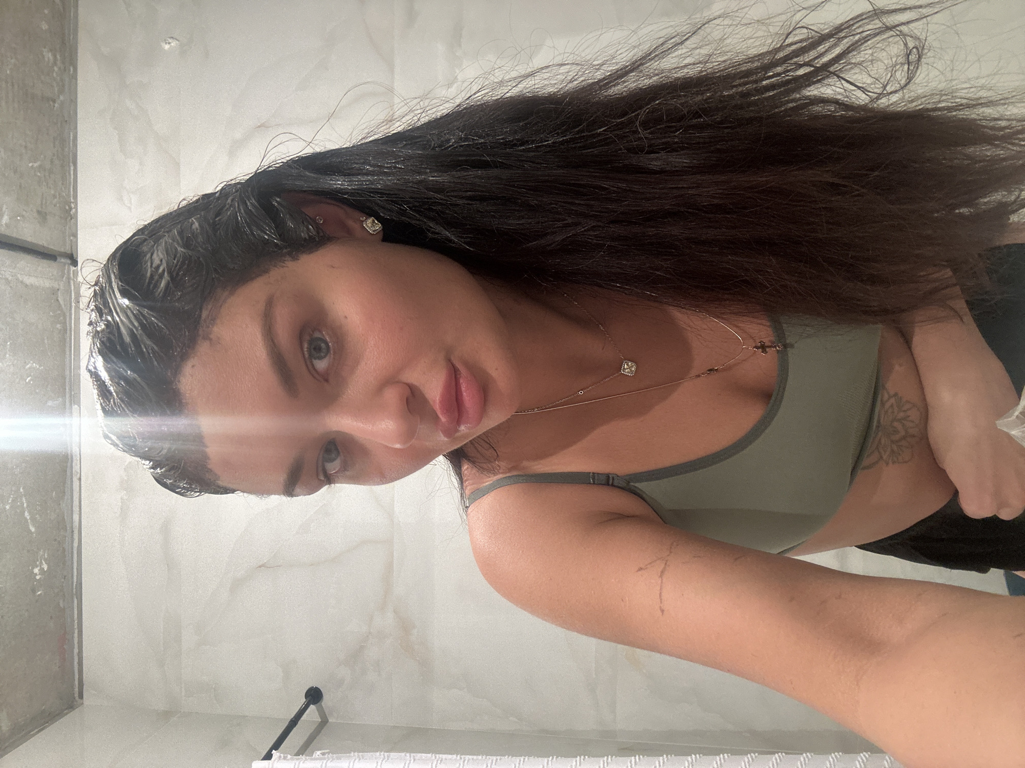 The image, rotated 90 degrees clockwise, captures a woman taking a self-portrait in what appears to be a bathroom. She is positioned horizontally across the frame, looking directly at the camera with a neutral expression and slightly parted lips. Her long, dark hair is spread out, with some strands appearing wet or damp around her forehead. She wears a form-fitting, olive-green sports bra or tank top, which reveals a portion of her torso and a floral-patterned tattoo on her left ribcage area. A delicate necklace with small, square, sparkling pendants adorns her neck. On her right arm, several faint, thin markings are visible.
The background consists of light-colored, possibly marble-tiled walls, indicative of an indoor setting like a modern bathroom. To the far left, a reflective surface, possibly a shower door or mirror, is partially visible. A dark-colored towel bar is also present in the lower left portion of the frame. A prominent lens flare from an artificial light source is visible on the left side of the image, near the woman's face, casting a bright illumination across that area. The overall scene is well-lit and appears clean. No visible text is present in the image. The woman's wet hair suggests she may have recently showered.
