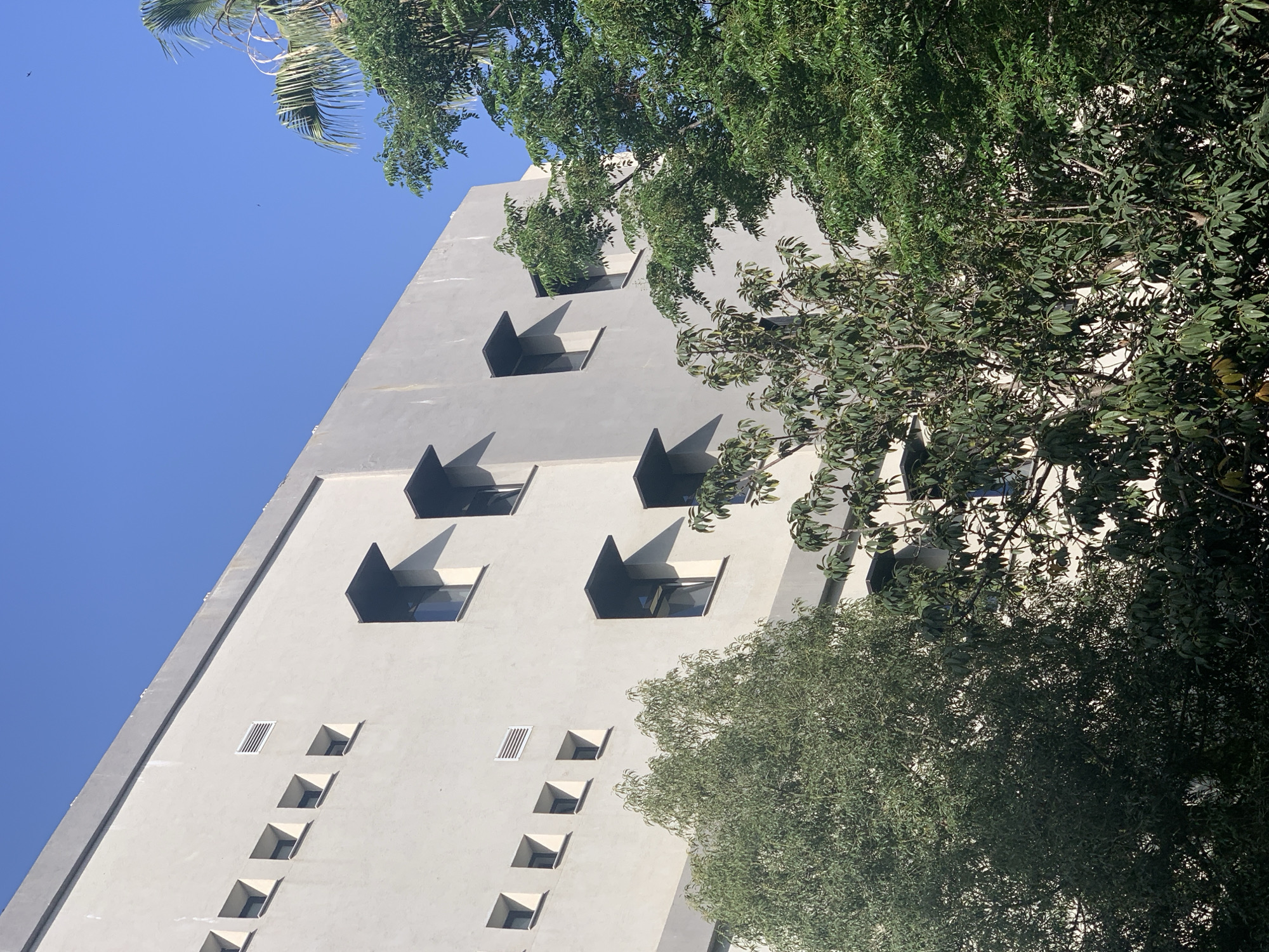 A low-angle shot reveals a tall, white building in Tel-Aviv, Israel, partially obscured by trees. The building features a unique architectural design with distinctive square windows and sunshade. Some windows are partially obscured by trees. The sky is blue and clear.