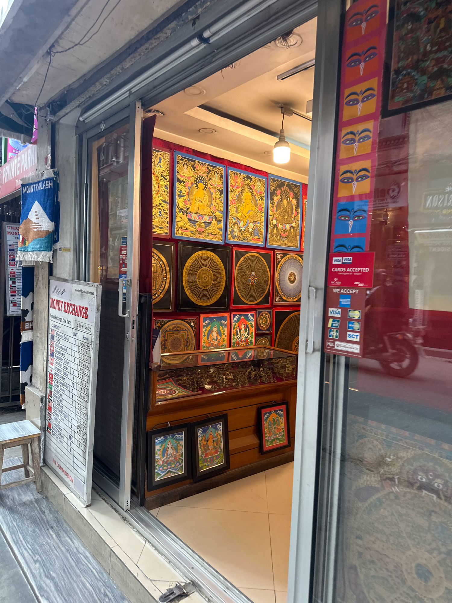 The image shows the exterior of a shop in Kathmandu Metropolitan City, Nepal, with a view into the interior. The shop appears to be a money exchange and souvenir store. In the foreground, a sign for "MONEY EXCHANGE" is visible, displaying current exchange rates for various currencies. There are also payment acceptance signs for major credit cards like Visa and Mastercard. The shop's interior is decorated with vibrant, traditional Nepalese paintings and artwork, possibly thangka paintings, featuring religious figures and mandalas. A display cabinet contains what appear to be small statues or religious artifacts. The exterior of the shop has a metal roller shutter and a glass door. A blue tapestry with the text "MOUNT KAILASH" is hanging on the left side of the shop entrance. The overall environment suggests a bustling commercial street.