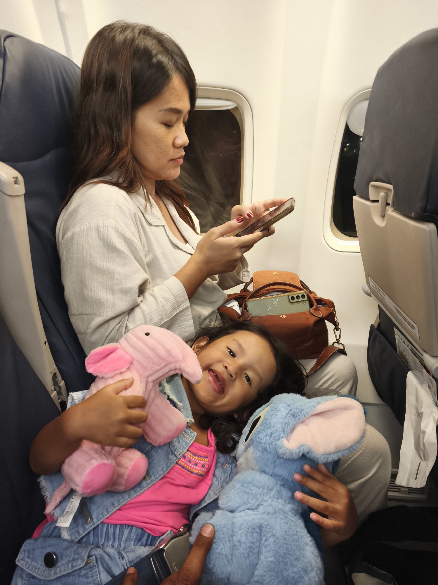 A young child is seated on an airplane, leaning against their mother. The child holds a pink elephant plush toy in their left arm and a blue stuffed animal, possibly Stitch, in their right arm. The child wears a denim jacket over a pink shirt and denim pants. The mother, seated beside the child, is looking at her phone. A brown handbag is visible between them. The scene is set within an airplane cabin, suggesting travel. There are no visible indicators that this location is in Kuta, Indonesia.