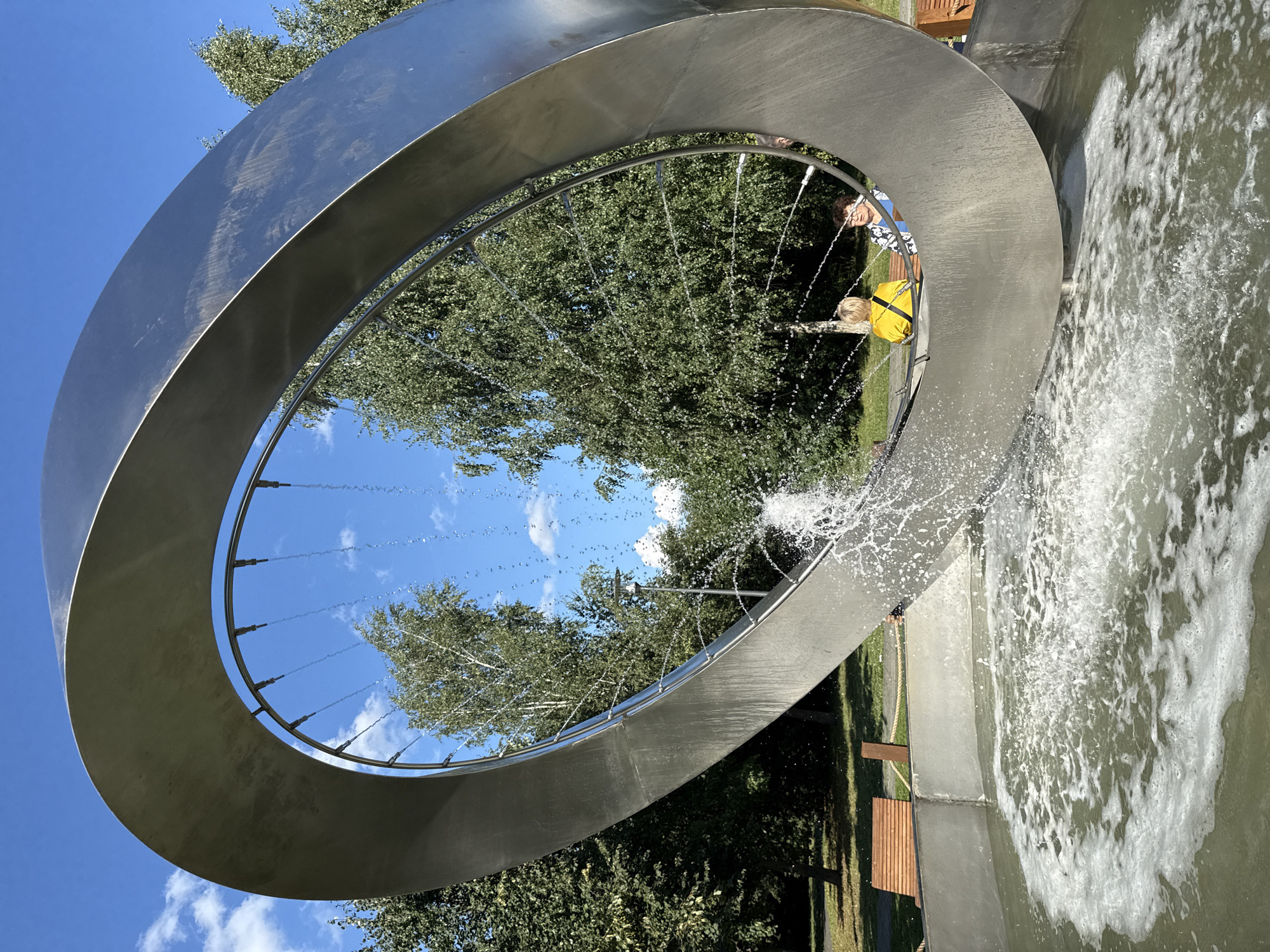 A large, oval, metal sculpture with an opening in the center is partially submerged in a fountain basin filled with rippling water. The sculpture is tilted, and water sprays from small nozzles arranged in a circular pattern around its inner edge, creating a cascading effect. Two individuals are seated on a bench within the opening of the sculpture, observing the fountain. A third person is partially visible in the background, also seated. The fountain is situated in a park-like setting with lush green trees and a bright blue sky with scattered clouds overhead. Wooden benches are visible along the edge of the fountain, and a paved pathway is present in the background. The sculpture appears to be a modern art installation located in the city of Khimki, Russia.