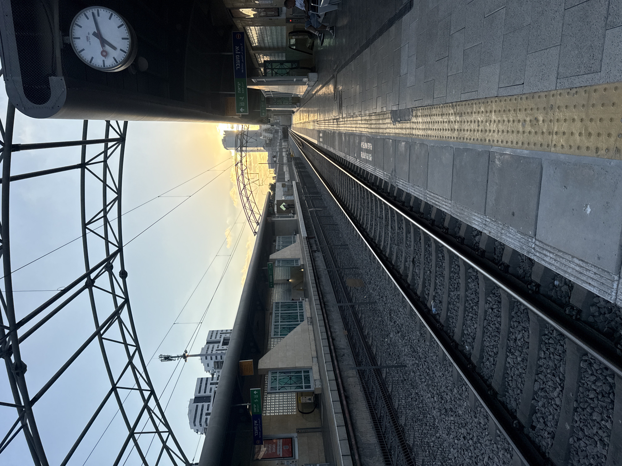 This is a photograph of the train station platform in Hod HaSharon, Israel, during sunset. The image is taken from the perspective of someone standing on the platform, looking down the train tracks. The tracks are visible in the foreground, with a train's electrical wires overhead.
On the right side of the image is the train platform, which is paved with gray tiles. A yellow tactile paving strip runs along the edge of the platform. A man is sitting on a bench on the platform. There are signs with Hebrew text and exit signs visible. A large clock is mounted on the underside of the station's roof.
On the left side of the image, across the tracks, is another platform with a covered waiting area and glass-paneled walls. In the background, tall, modern buildings can be seen, illuminated by the setting sun, which creates a warm glow in the sky. The sky is a pale blue with some clouds. The overall scene depicts a quiet moment at a public transportation hub as the day ends.