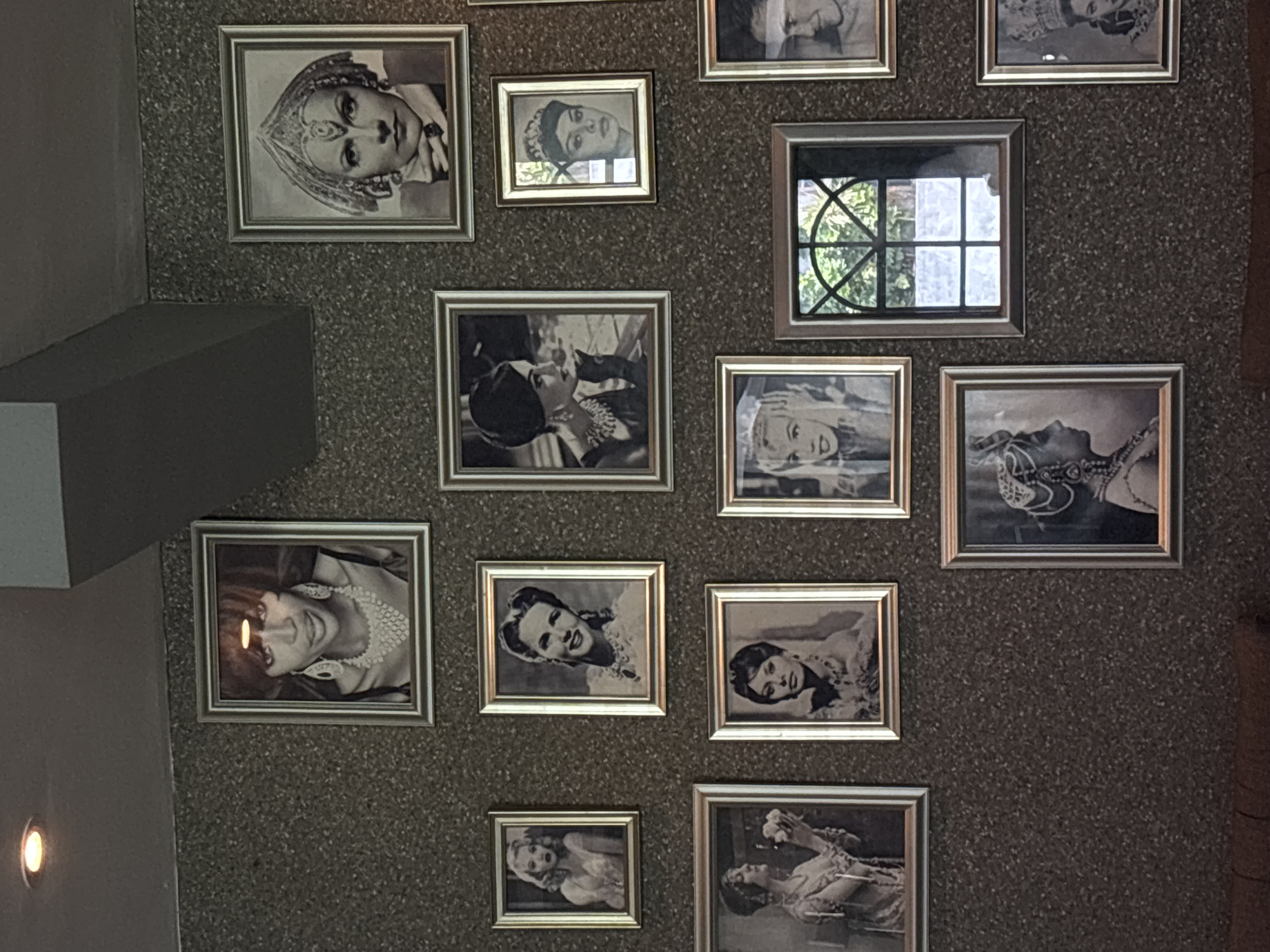 A wall in North Kuta, Indonesia, is adorned with framed black-and-white photographs. The frames are a uniform metallic color, possibly silver or gold. The photos predominantly feature portraits of women, some wearing ornate headwear or jewelry. A single framed window reflects an outdoor scene with green foliage and a grid pattern. The wall has a textured, speckled surface. A gray structural element and a light fixture are visible on the left side of the frame.