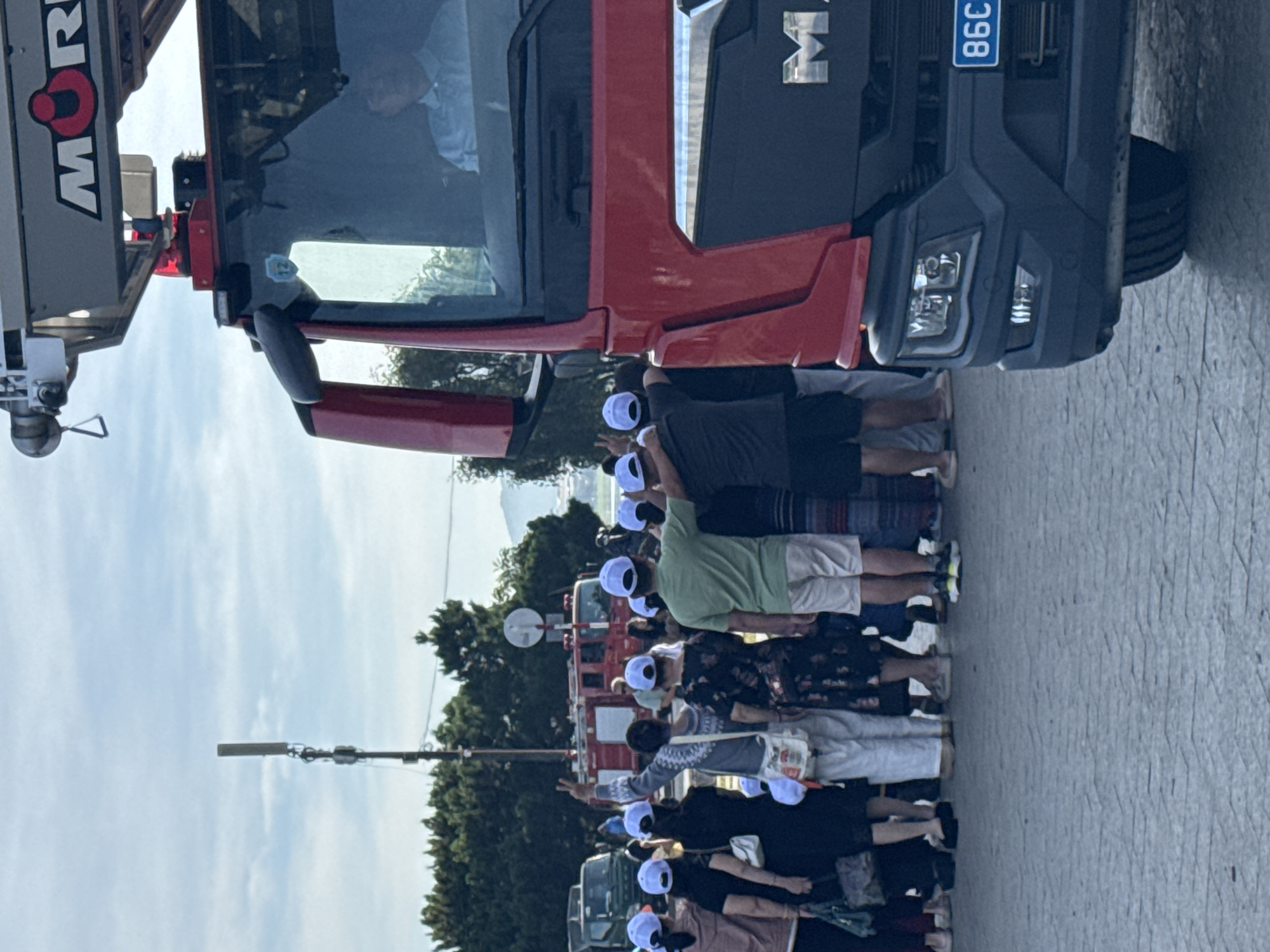 In Da Lat, Vietnam, a red fire truck is parked on a paved surface, possibly a street or parking lot, with a group of people gathered in front of it. The fire truck has "MOR" emblazoned on its side. A man in a light blue collared shirt is visible through the windshield, appearing to be the driver. The crowd, mostly wearing white baseball caps, faces away from the camera, with some individuals raising their hands. Trees and a utility pole are visible in the background under a cloudy sky. License plate reads "86C".