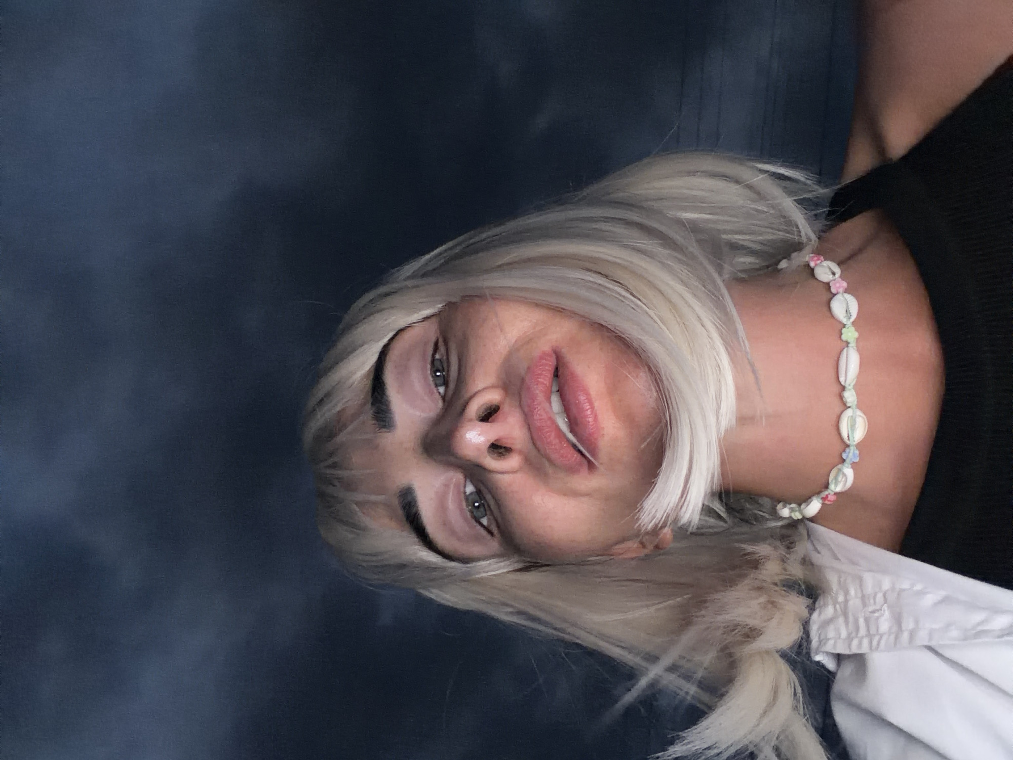 A low-angle selfie shows a woman with blonde hair and bangs, wearing a shell necklace and a black tank top. Her face is turned towards the camera, with her mouth slightly open. The background is a dark, cloudy sky, suggestive of an outdoor nighttime setting in Sochi, Russia.