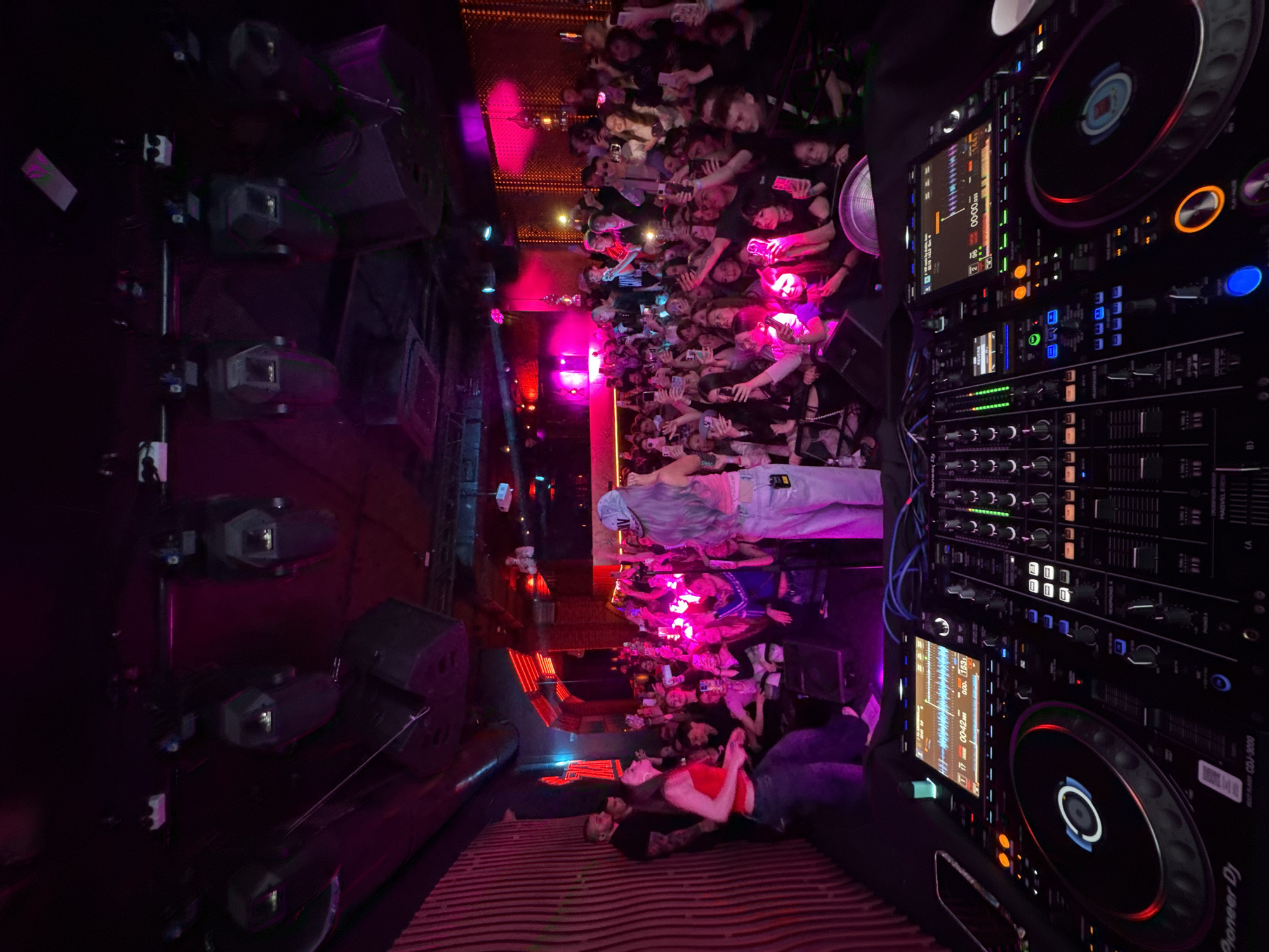 This media file captures a vibrant, high-angle view of a nightclub or concert event in Khabarovsk, Russia, taken from an elevated position looking down. The image is rotated 90 degrees counter-clockwise, showing the scene vertically.
On the right side of the frame, a professional DJ setup is prominently displayed, featuring Pioneer CDJ-3000 decks and a mixer with numerous illuminated buttons and screens. Visible text on the equipment includes "Pioneer DJ", "CDJ-3000", and digital readouts like "000. 1531" and "00:00.033". Blue and red cables connect various components.
To the left of the DJ equipment, a large crowd of people fills the middle ground, stretching towards the left edge of the frame. Many attendees are holding up their smartphones, presumably recording or taking pictures of the performance. The crowd is illuminated by intense pink and purple stage lights, creating a dynamic atmosphere.
Further to the left, a series of large, dark speakers or stage monitors are stacked vertically. In the lower left foreground, close to the DJ booth, two individuals are lying on a dark, horizontally striped surface, appearing relaxed or resting, with their heads facing away from the DJ equipment.
The overall scene conveys a lively night event, characterized by music, a large engaged audience, and dramatic lighting.