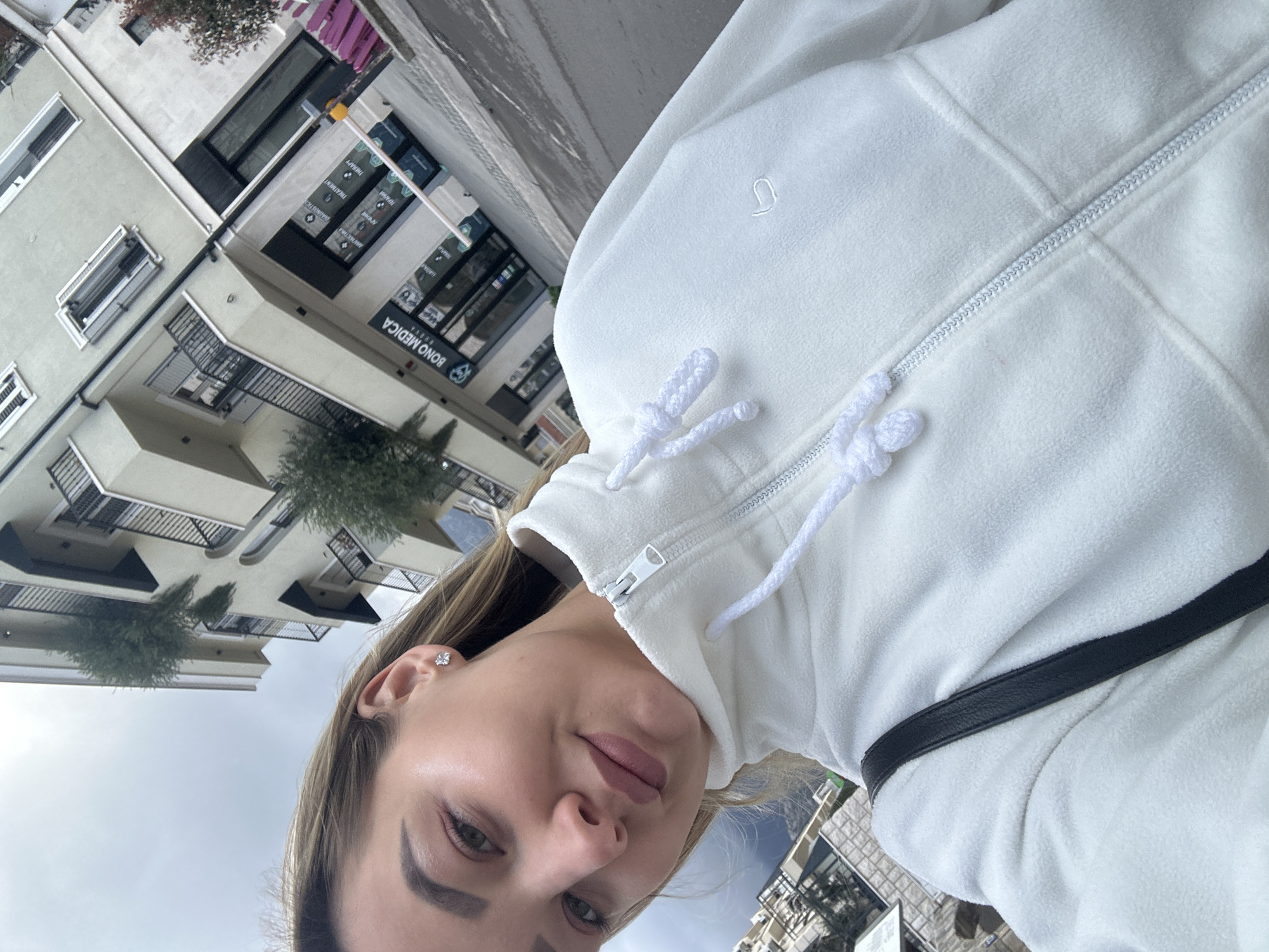 The image displays a close-up selfie of a light-skinned female individual, estimated to be between 20 and 30 years old, with light brown hair parted on her right side and light makeup, including defined eyebrows and pink lipstick. She looks directly at the camera with a slight pout. She wears a white fleece full-zip jacket with a high collar and white drawstrings. A small, white embroidered logo, resembling a stylized 'J' or 'U', is visible on the jacket's left chest. A black strap, consistent with a shoulder bag, crosses over her right shoulder. A small, clear, bow-shaped stud earring is present in her right ear.
The background shows an urban street scene under an overcast sky. Behind the individual, a multi-story building with a light-colored (beige/light grey) facade is visible, featuring multiple balconies with dark railings and some green foliage on the terraces. The ground floor of this building houses a business identified by the sign "BONO MEDICA BUDVA" on its glass storefront windows. Additional signage in the windows, partly in Cyrillic and English, advertises services such as "DIAGNOSTICS," "TREATMENT," and "THERAPY." A white and red barrier arm, typically used for vehicle access control, is partially visible in front of "BONO MEDICA." The street surface in the immediate foreground appears damp. The scene is specifically located in Budva, Montenegro, in front of the Bono Medica polyclinic.