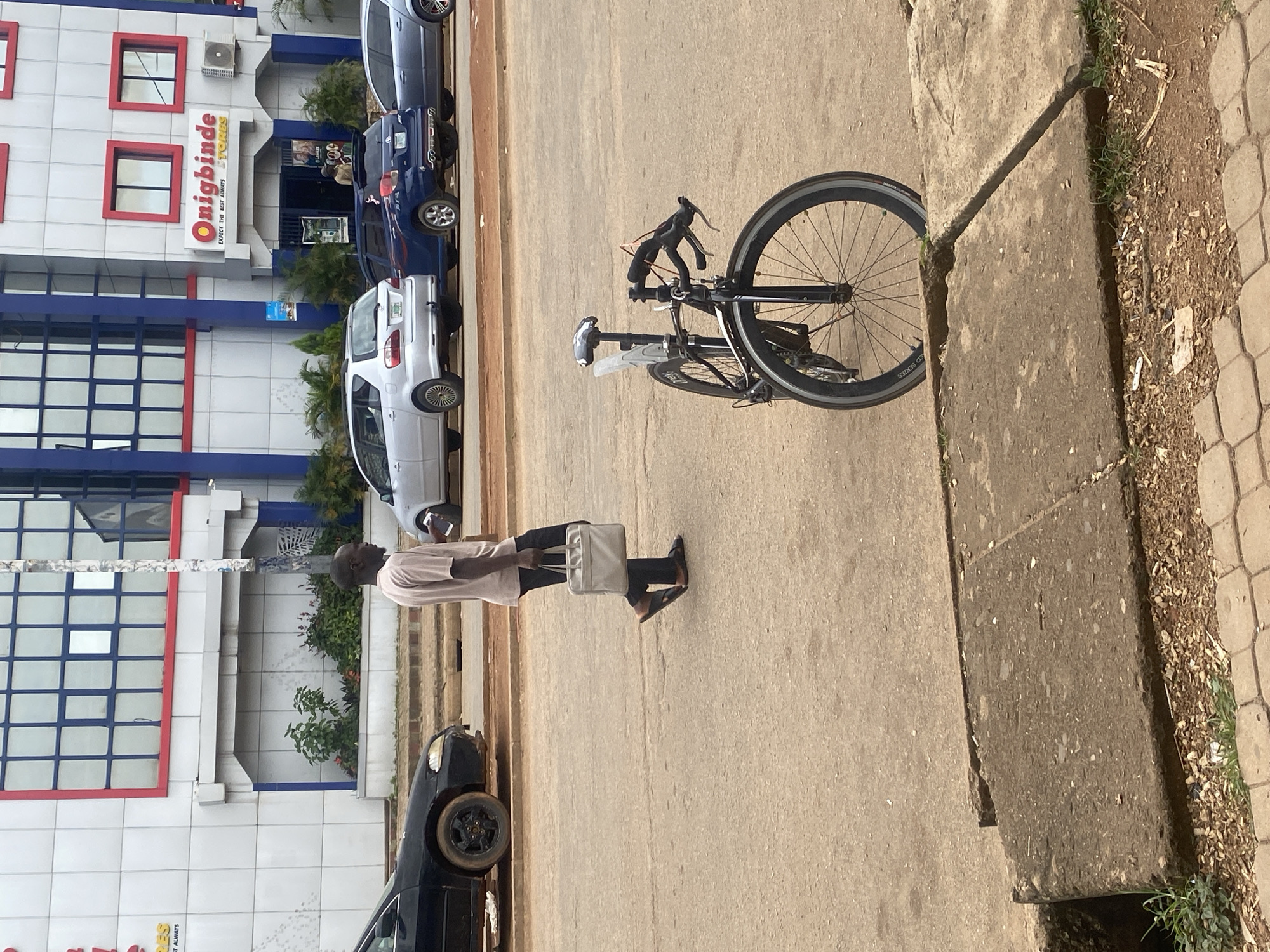 A man walks on a paved street carrying a light-colored bag in his right hand and a mobile phone in his left. He is wearing a light-colored short-sleeved shirt and dark trousers. In the foreground, a black bicycle is parked on the street, leaning on its kickstand. Behind the man, several cars are parked in front of a multi-story building with a sign that reads "Onigbinde STORES." The building has a modern architectural design with blue and red accents. The street appears to be in a commercial area.
