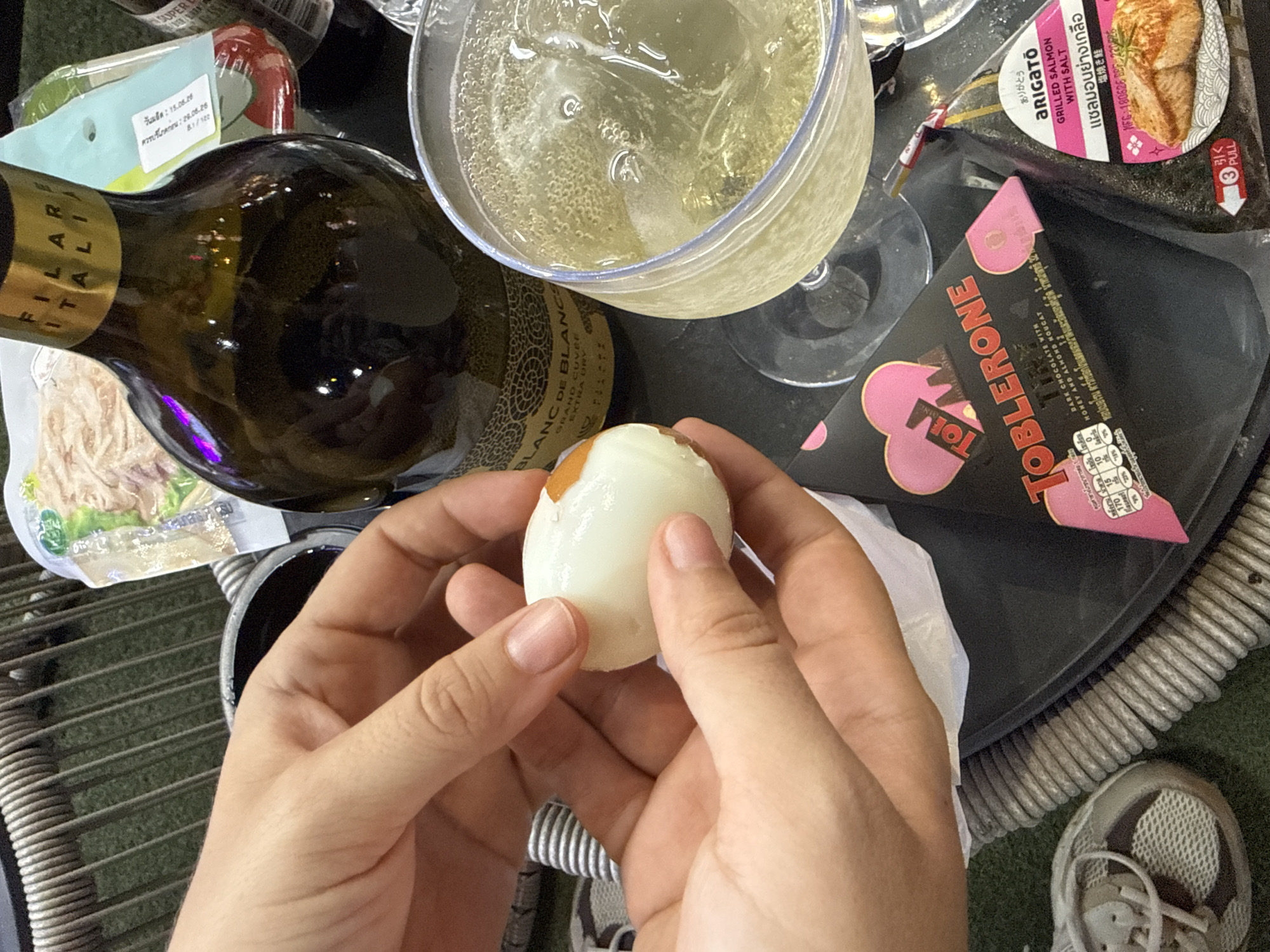 This is a close-up, top-down shot of a person holding a peeled hard-boiled egg over a dark, textured surface, possibly a tray or table. The setting appears to be casual, with the edge of a woven chair visible in the foreground.
On the surface, several items are arranged around the egg. To the left, a dark green bottle with a gold label partially obscures a clear glass of bubbly, light-colored liquid, likely champagne or sparkling wine. The bottle's label reads "FILARE ITALIA" and "BLANC DE BLANCS GRAND CUVEE EXTRA DRY". Further left, a white and green package is visible, with Thai text on a light blue background.
To the right of the egg, a triangular package with pink accents is prominent, displaying the brand name "TOBLERONE" in large letters. Next to it, a round package features an image of grilled salmon and has text in multiple languages, including "ARIGATO" and "GRILLED SALMON WITH SALT". Thai text on this package includes "ควรบริโภคก่อน : 28.06.26" (Consume before: 28.06.26) and "วันมด : 15.08.26" (Manufacturing date: 15.08.26). There is also a "PULL" indicator on this package.
The lighting suggests it could be daytime, possibly indoors or in a shaded outdoor area. The focus is on the hands holding the egg and the surrounding items, giving a sense of an impromptu snack or meal. The overall mood is relaxed and informal. The shoes visible at the bottom right are athletic sneakers, further reinforcing a casual setting.