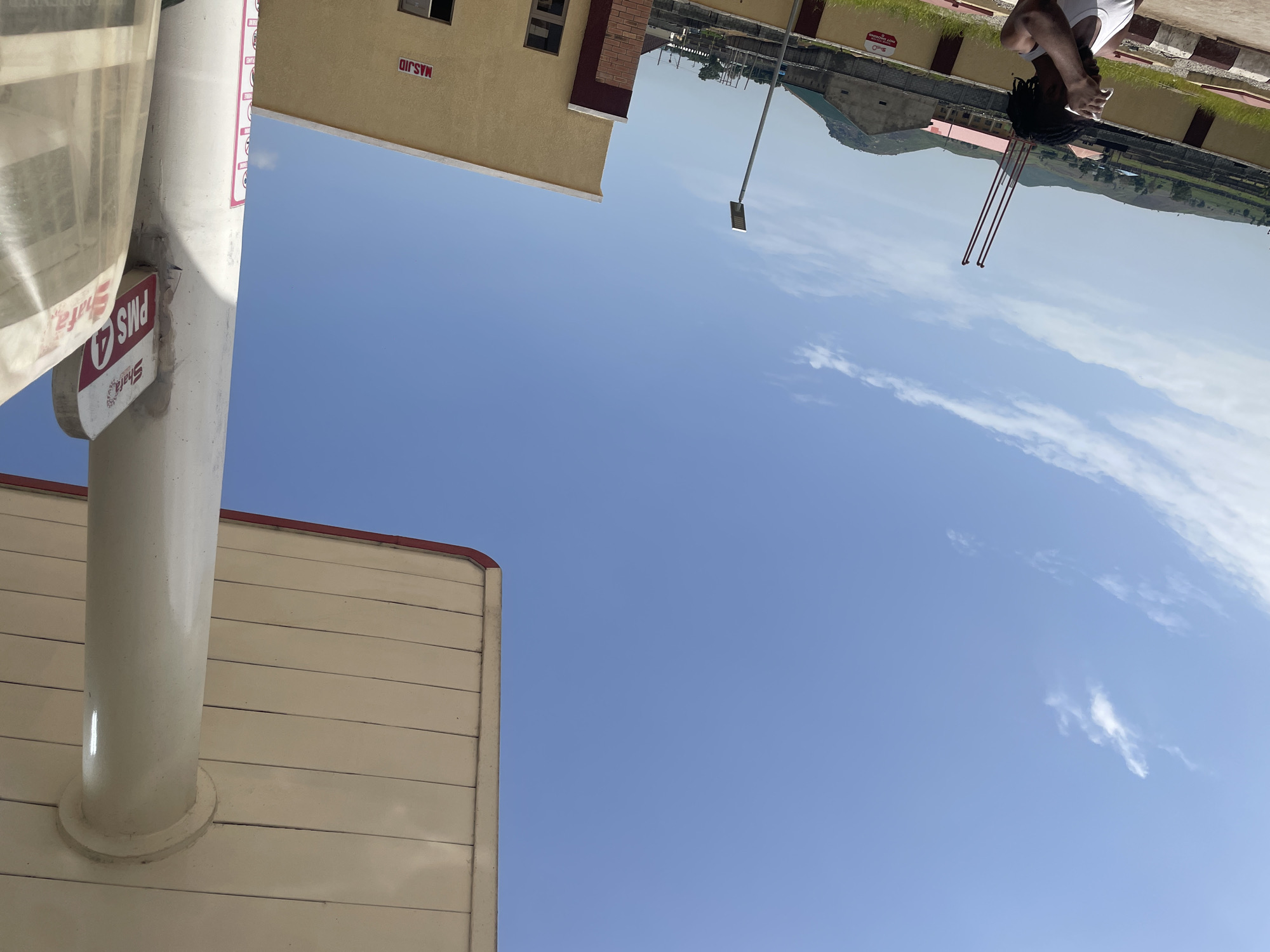 A person with dark dreadlocks, wearing a white tank top, stands at a gas station in Pasere, Nigeria, wiping their forehead. The sky is clear blue with scattered white clouds. A concrete pillar supports the roof of the gas station canopy, which has a beige ceiling and a red trim. A sign on the pillar reads "Shafa PMS 4". In the background, a light yellow building with a red trim has the word "MASJID" written on its exterior. Other buildings and structures, including some with red roofs, are visible further in the distance, along with rolling green hills. The ground appears to be paved with some areas of grass and concrete barriers. A street light stands in the middle ground.