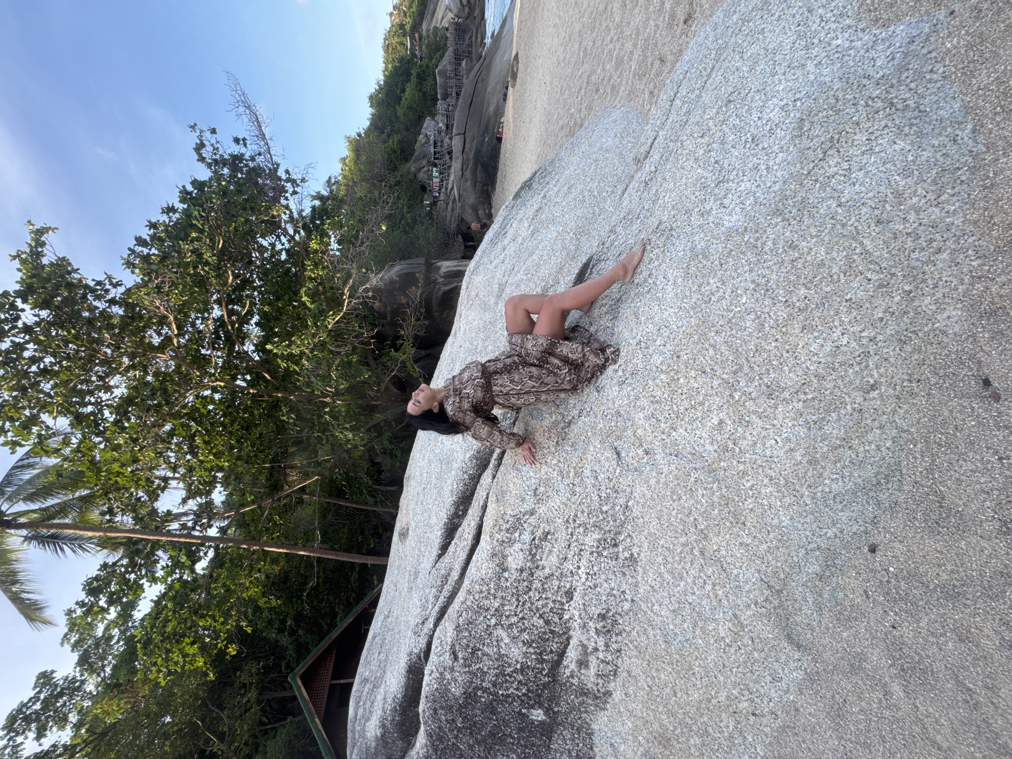 A woman is lying on a large rock on a beach in Ban Haad Rin, Thailand. She is wearing a long-sleeved, patterned dress. Her head is tilted back, and she is looking upward. She is barefoot. The rock is large and gray. There are trees and vegetation surrounding the rock. The sky is blue and mostly clear. In the background, there is a beach with some structures.