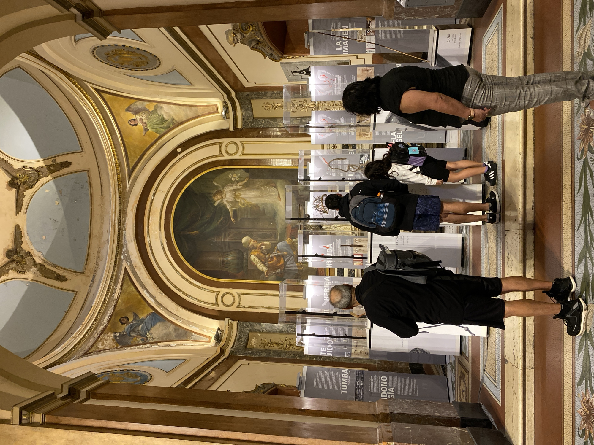 The image captures an interior scene within a grand, ornately decorated building, likely a museum or a historical site, in Buenos Aires, Argentina. The setting features elaborate architecture, including arches, intricate moldings, and painted murals on the ceiling and upper walls, depicting angels and other figures. A prominent large painting, possibly an altarpiece with a historical or biblical theme, is visible on the far wall. The floor is adorned with decorative tiles.
Four people, consisting of adults and one child or young person, are actively observing an exhibition. They are positioned in front of several rows of vertical, transparent display cases, which contain informational panels, images, and possibly artifacts. The individuals are seen from behind or the side, appearing focused and engaged with the exhibits. One of the display cases clearly shows what appears to be a replica or representation of a Crown of Thorns, hinting at the exhibition's religious subject matter.
Visible text on the display panels, confirmed by OCR, includes titles such as "LA IMAGEN" (The Image), "FLA GEL" (Flagellum), "HERIDA" (Wound), "TE JIDO" (Fabric/Tissue), "TUMBA" (Tomb), and "CAÑA" (Cane/Reed). A biblical reference, "Mateo 27:29-30" (Matthew 27:29-30), is also present, reinforcing the religious context, possibly related to the Passion of Christ. The lighting suggests an indoor daytime setting, illuminated by a combination of natural and artificial light. The atmosphere appears to be one of quiet contemplation and cultural engagement.