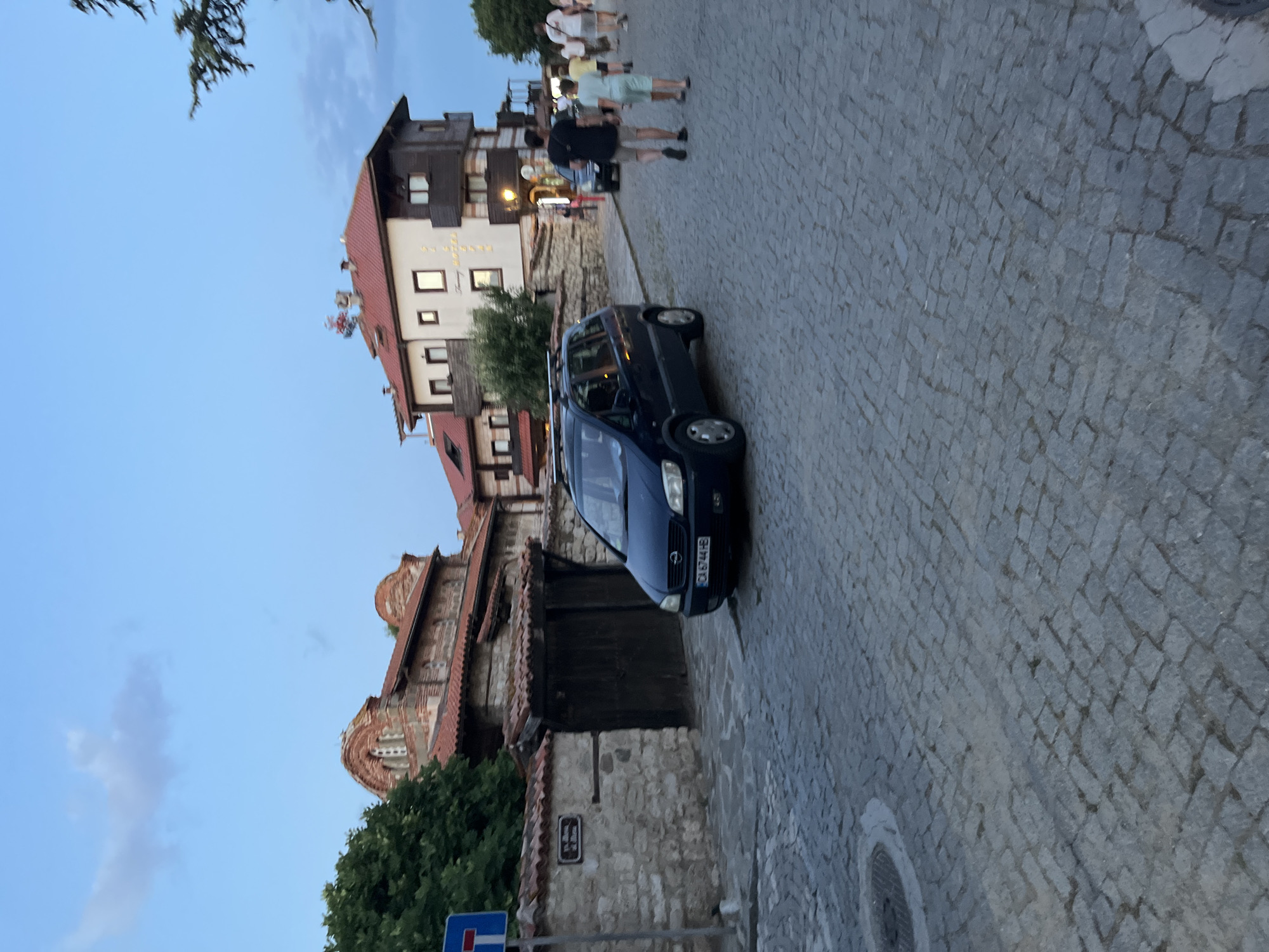 The image shows a street scene in Nesebar, Bulgaria, likely during the evening. Cobblestones pave the street and sidewalk. On the left, a stone wall encloses a courtyard with a dark wooden gate. Above the wall, red-tiled roofs of ancient buildings, possibly churches, are visible, constructed with exposed brick and arched windows. A dark blue minivan is parked on the street in front of the gate, with its license plate "CA 6744 HB" partially visible.
Further down the street, on the right, a multi-story building with a prominent red-tiled roof and dark wooden accents stands. This building appears to be a hotel, with signage that reads "Sunny Hotel". Several people are walking along the street, some heading towards the hotel. Among them, a man in a black t-shirt and shorts walks with a younger person in a light blue t-shirt and shorts. Others are visible further in the background, also walking. The sky above is a pale blue, indicating twilight. Trees and branches are visible on the far right of the image. A blue traffic sign with a white horizontal bar and a red vertical bar is present on a pole to the left of the scene. A circular manhole cover is visible in the cobblestones in the lower left of the image.