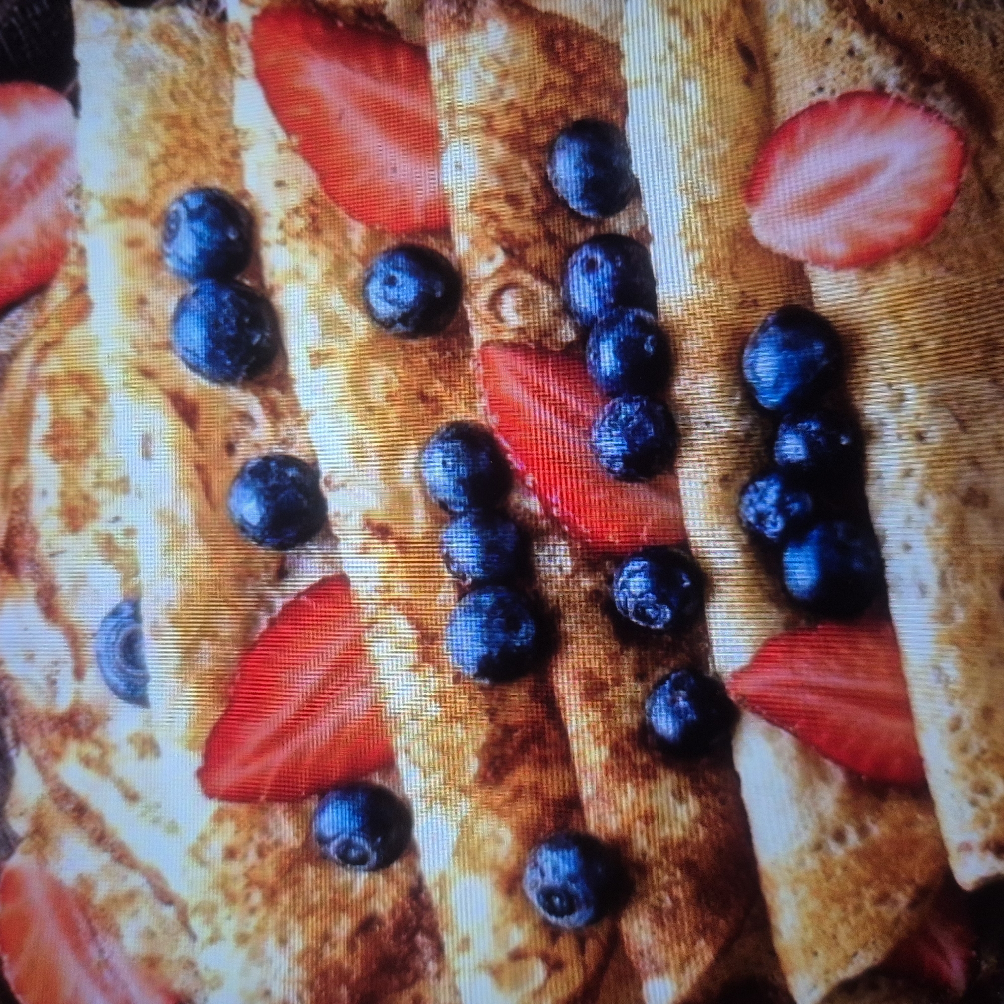 The image shows a close-up, overhead view of several rolled crepes, likely crepes or thin pancakes, arranged in a layered fashion. Scattered on top of and between the crepes are fresh blueberries and slices of strawberries. The crepes appear golden brown and slightly textured, suggesting they have been cooked. The lighting highlights the glossy sheen of the berries and the soft folds of the crepes. There are no people or explicit environmental details visible that would indicate a specific city or location like Nowa Wieś Książęca, Poland. The scene is solely focused on the food presentation.