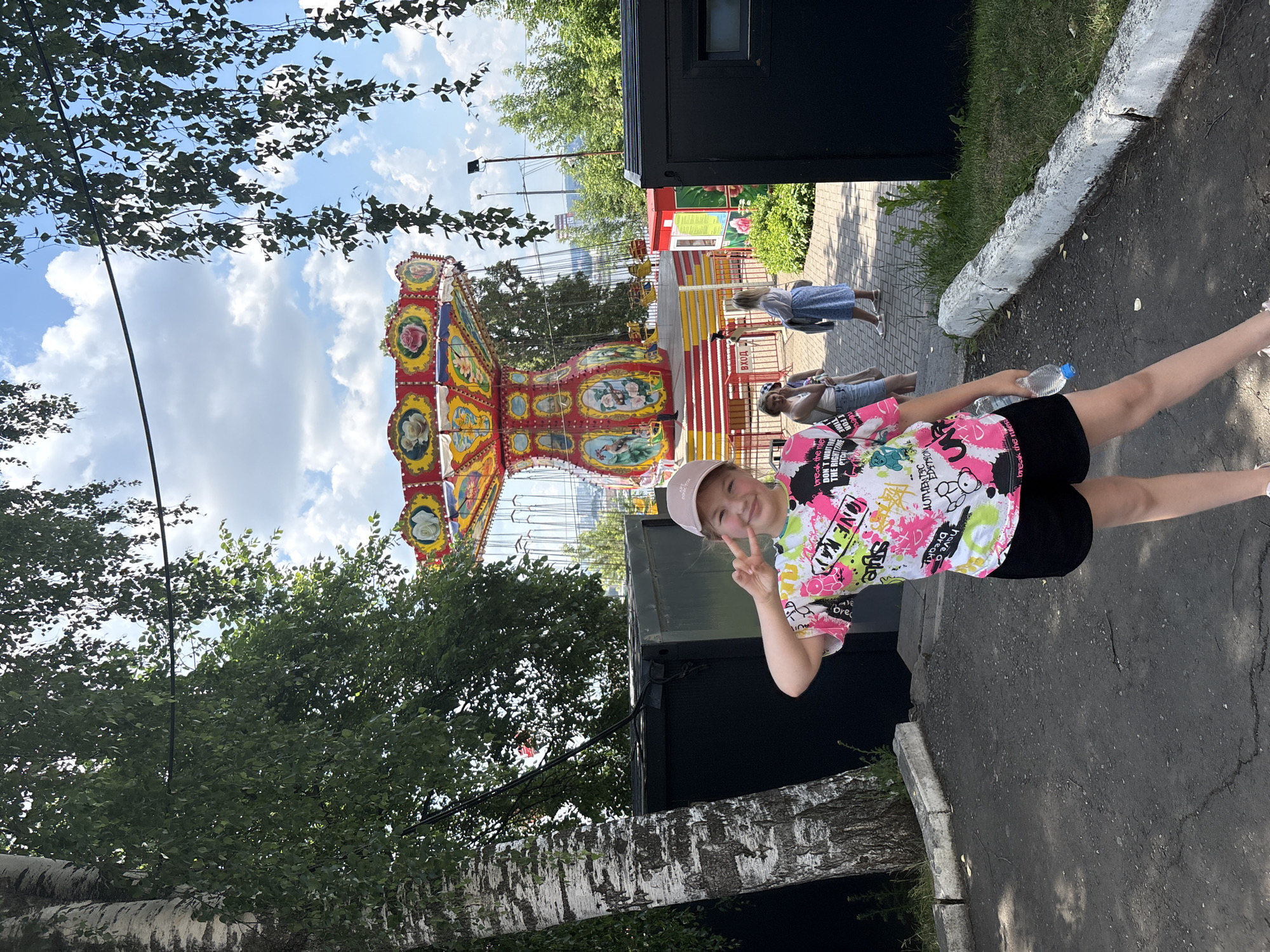 The image depicts a young girl wearing a colorful t-shirt and black shorts, standing in an outdoor amusement park area. She is smiling and making a peace sign with her fingers. Behind her, a swing ride attraction is visible, along with trees and a cloudy sky. Several other people are in the background, some walking and others near the ride. The ground is paved with asphalt and there is grass on the sides. The girl is holding a water bottle in her right hand. The location appears to be a part of the Izhevsk Zoo, specifically near the "Attractions" area, a popular spot for families in the city of Izhevsk, Russia.