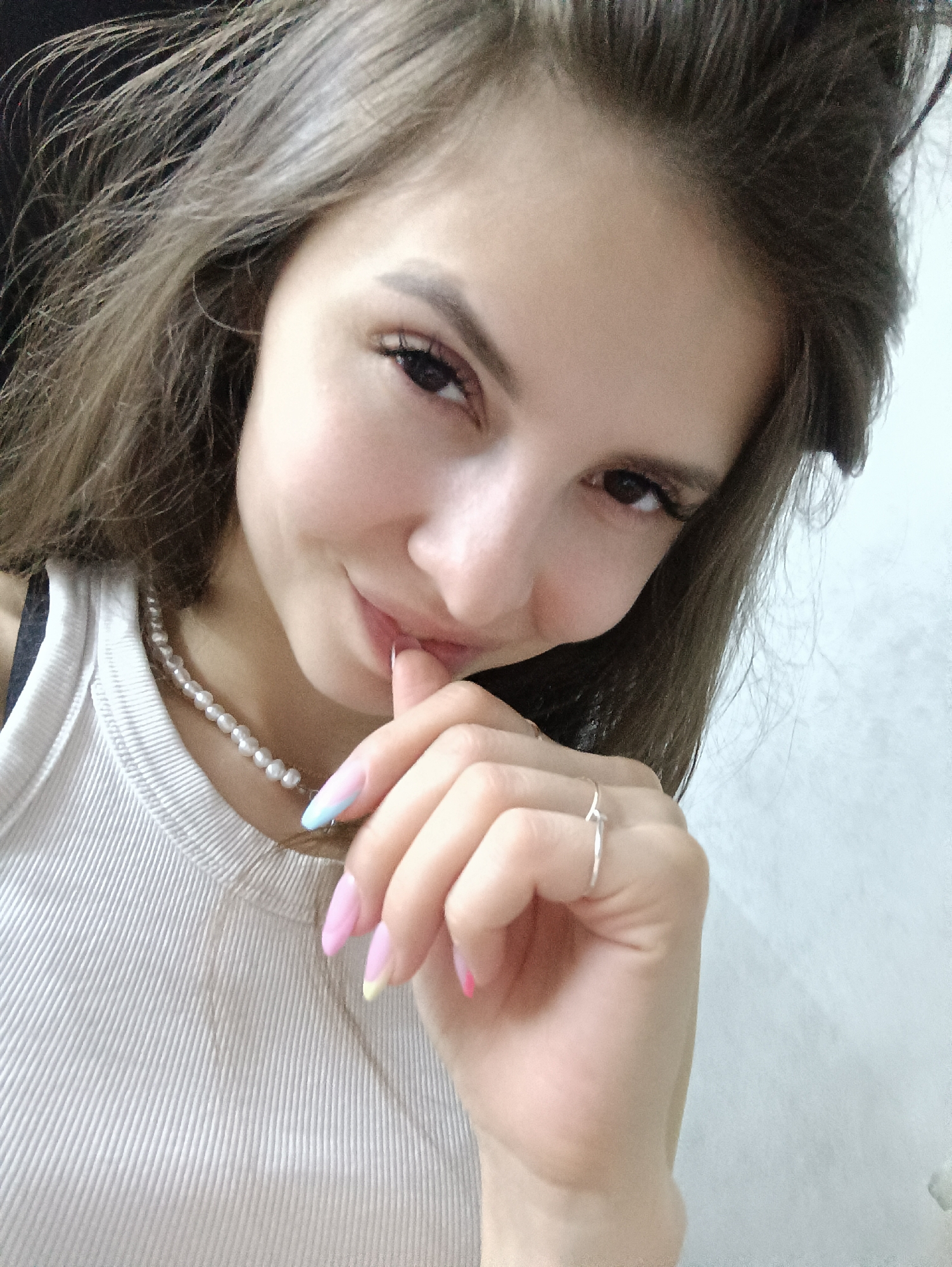 A young woman is photographed in a close-up selfie shot. She has long, wavy brown hair, dark eyes, and fair skin. She is wearing a white ribbed tank top and a pearl necklace. Her nails are painted in a pastel color palette. A silver ring is visible on her finger, which is held to her lips. The background is out of focus, revealing a dark area on the left and a light-colored wall on the right. The photograph was taken in the city of Chaikovsky, Russia.