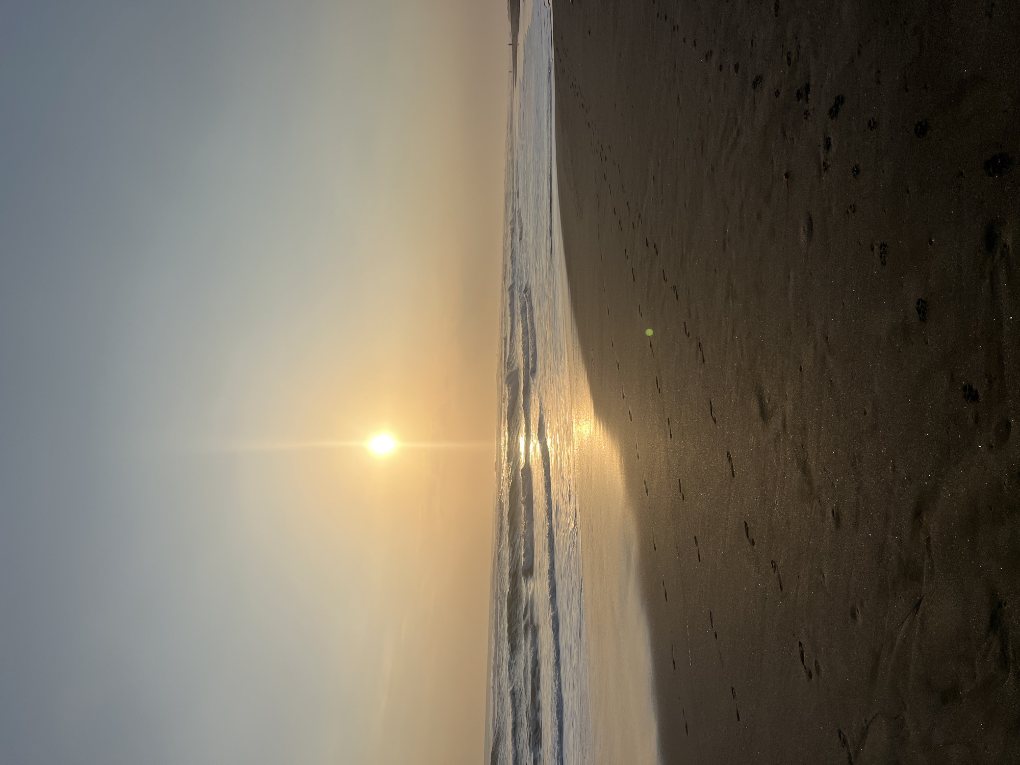 A wide shot of a sandy beach with footprints leading across the foreground. The sun is low in the sky, casting a warm glow on the scene and reflecting on the ocean waves. The sky is a gradient of pale yellow to light blue. There is a single person visible in the distance on the right side of the frame, walking along the shoreline. The image does not provide specific details about the city of Kediri, Indonesia, beyond its potential geographic setting as a coastal area.