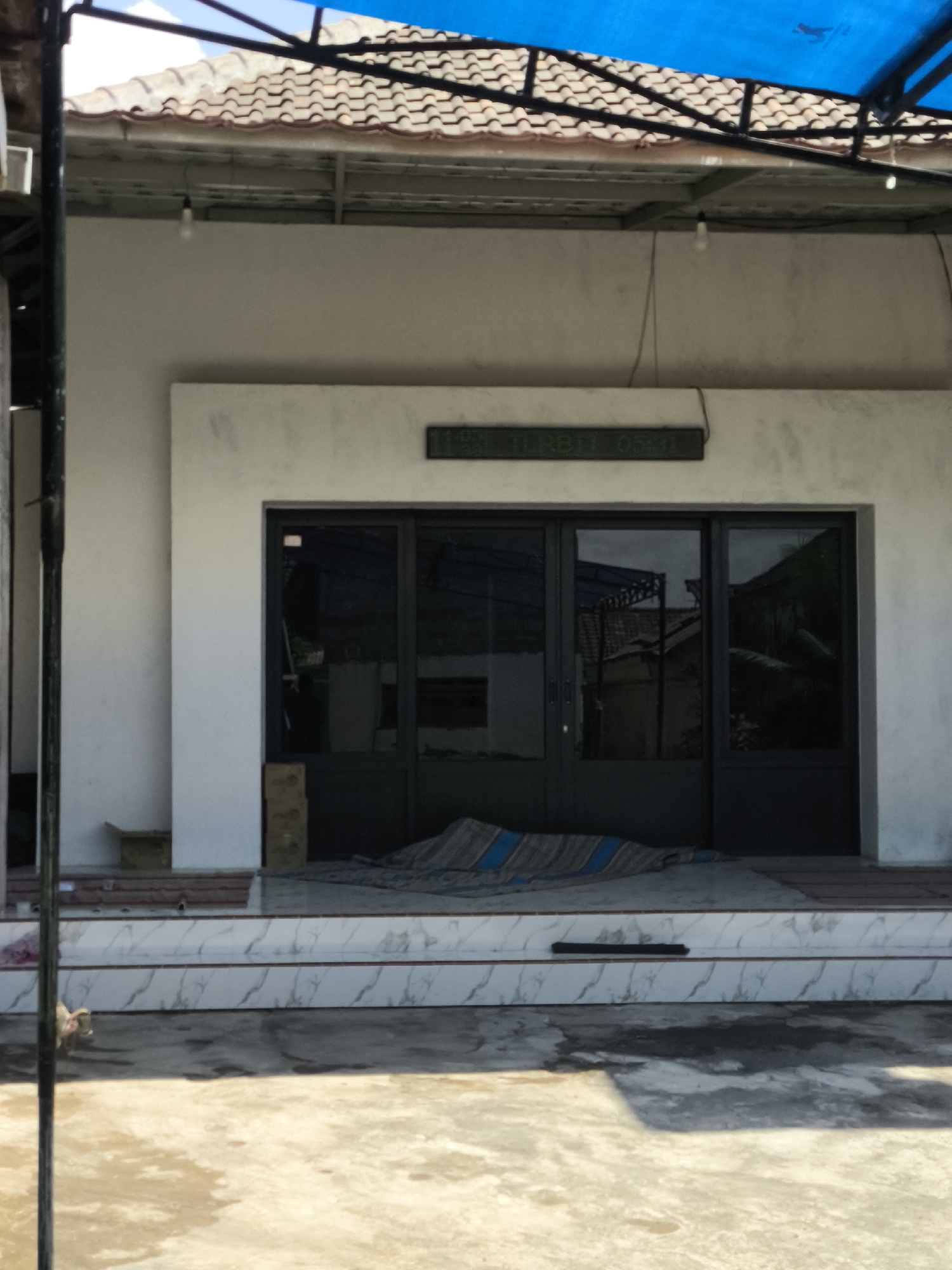 The image depicts the exterior of a building with a light-colored facade, featuring a set of dark-framed glass doors. Above the doors, a digital display shows the text "12.11 RABU 05:31". The building is partially covered by a brown tiled roof, and a blue tarp is visible overhead, supported by black metal framing. In front of the doors, there are steps with white marble-like tiles. A striped rug or mat is placed on the steps. The ground in front of the building is a paved area with visible water stains and shadows. To the left of the building, a dark vertical pole is present. The scene appears to be an outdoor, daytime setting, possibly a shop or commercial establishment in Kebonsari, Indonesia.