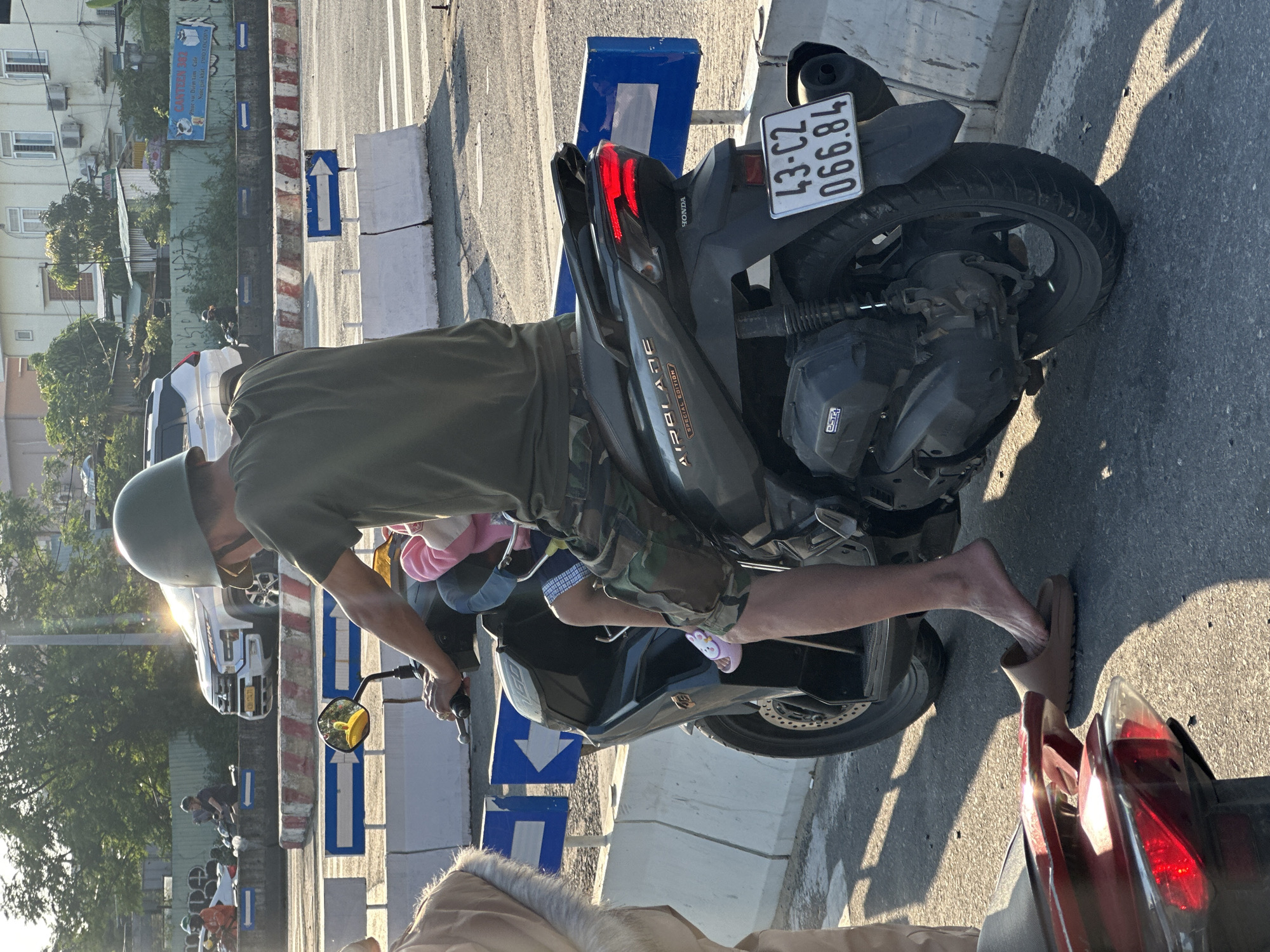 A man wearing a helmet, a green shirt, and camouflage shorts is riding a black Honda Airblade scooter in Đà Nẵng, Vietnam. The scooter's license plate number is 43-C2 066.84. A child wearing a pink outfit sits in front of him. The man is wearing a tan sandal on one foot and is barefoot on the other. The scooter is on a street with road dividers and directional arrows, with buildings and trees visible in the background. A cafe sign is also visible that reads "Vu Diem Lam-Cafe".
