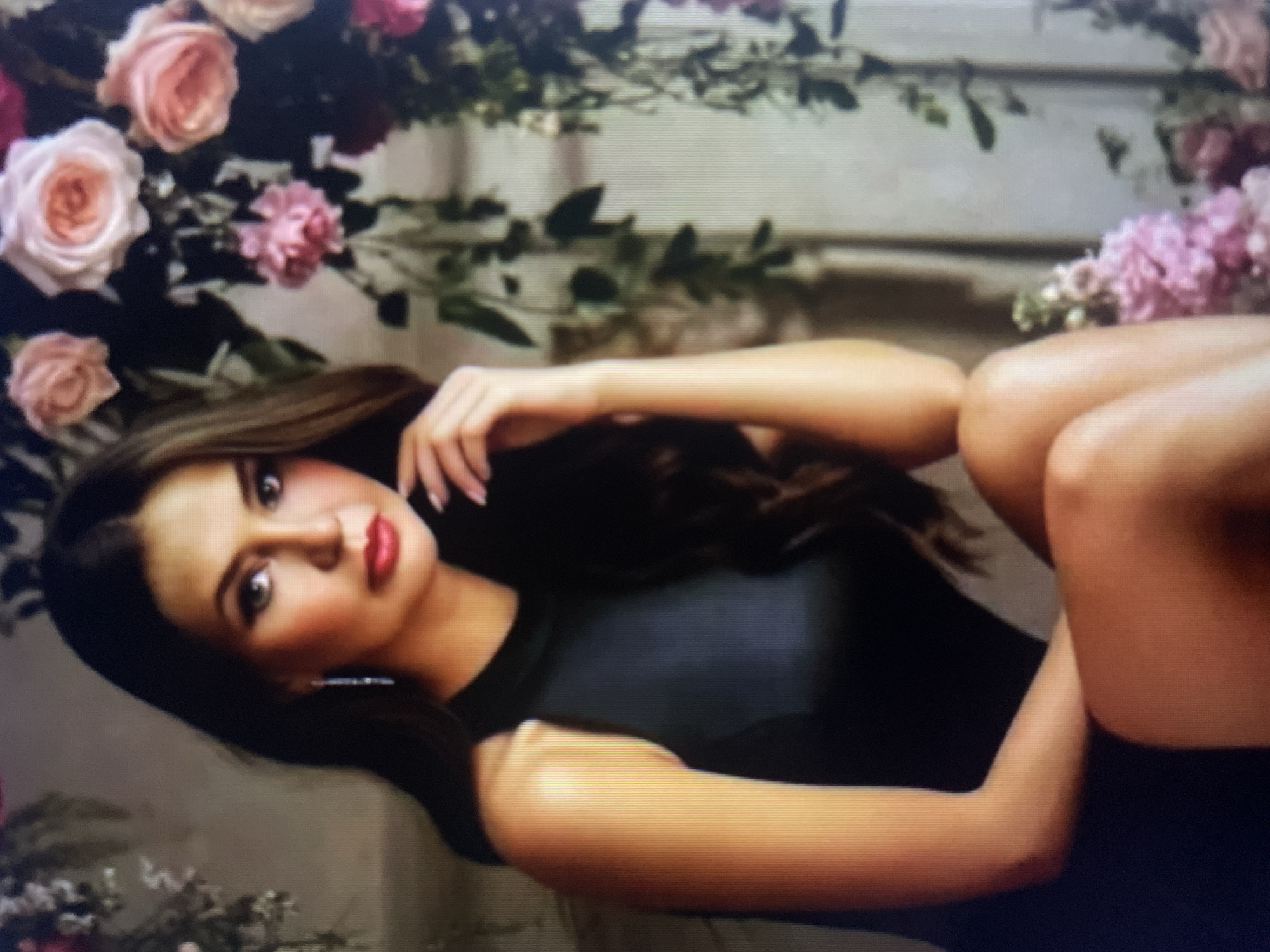 A woman with long dark hair and fair skin is depicted in a studio portrait, seated and looking towards the viewer. She is wearing a sleeveless black dress and delicate dangling earrings. Her left hand rests on her chin, and her right arm is bent at the elbow, with her hand near her face. The background features an abundance of pink and cream roses and green foliage, suggesting a floral arrangement or backdrop. The lighting is soft, highlighting her features and the textures of the flowers. There is no indication of the city of Kołobrzeg, Poland within the image.