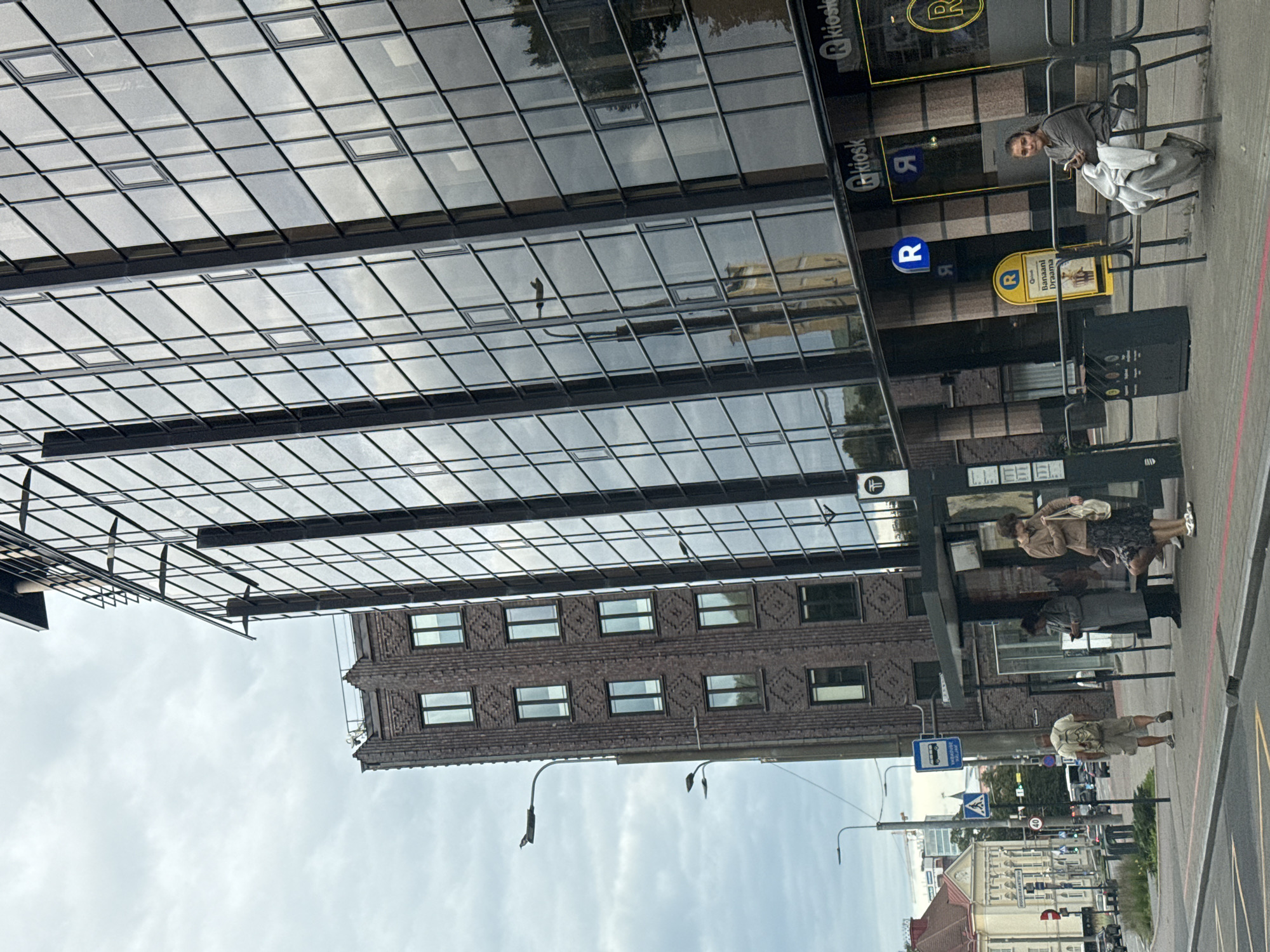 The image shows a street scene in Tallinn, Estonia. In the foreground, a bus stop shelter is visible with several people waiting. One woman is sitting on a bench, looking at her phone. Another woman stands next to her, holding a bag. A man with a backpack is walking away from the bus stop towards the street.
Behind the bus stop, a modern building with a glass facade dominates the scene. Reflections of the sky and nearby buildings are visible in the glass. To the left of the glass building is a tall, older brick building with a distinctive patterned facade. Streetlights and traffic signs are also present.
In the background, a street with cars and pedestrians can be seen, as well as other buildings characteristic of a city environment. The sky is overcast. The overall impression is of an urban setting on a cloudy day.