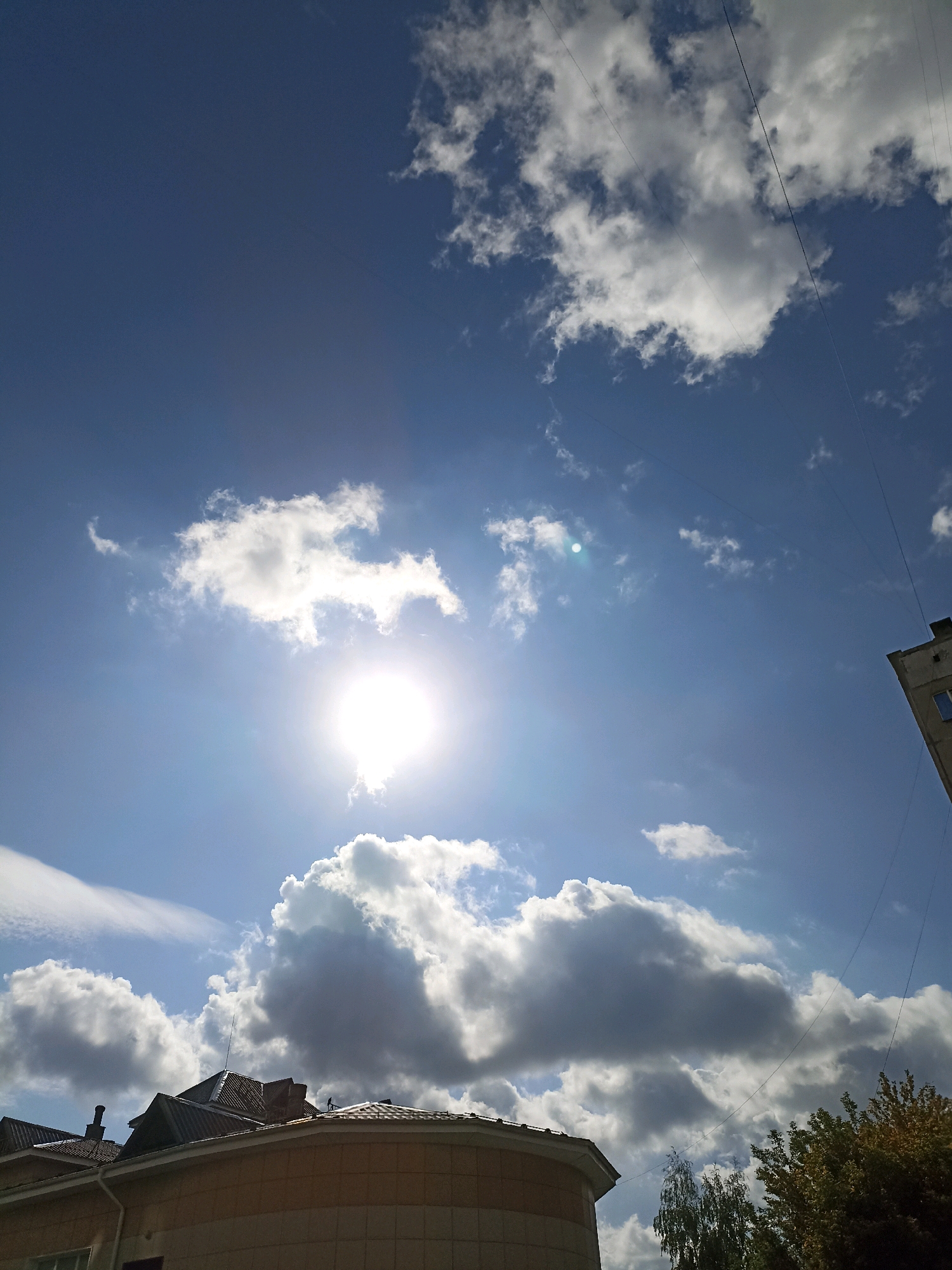 A bright, luminous sun is partially obscured by scattered white and grey clouds in a vibrant blue sky. Thin, dark wires traverse the sky from the upper right corner, and a portion of a gray building with windows is visible on the far right. In the lower portion of the image, the roofs of several buildings are visible, with one prominently featuring a beige facade and dark, angled rooflines. Trees with green and yellow foliage are also present in the lower right. The overall scene depicts an outdoor urban environment on a partly cloudy day.