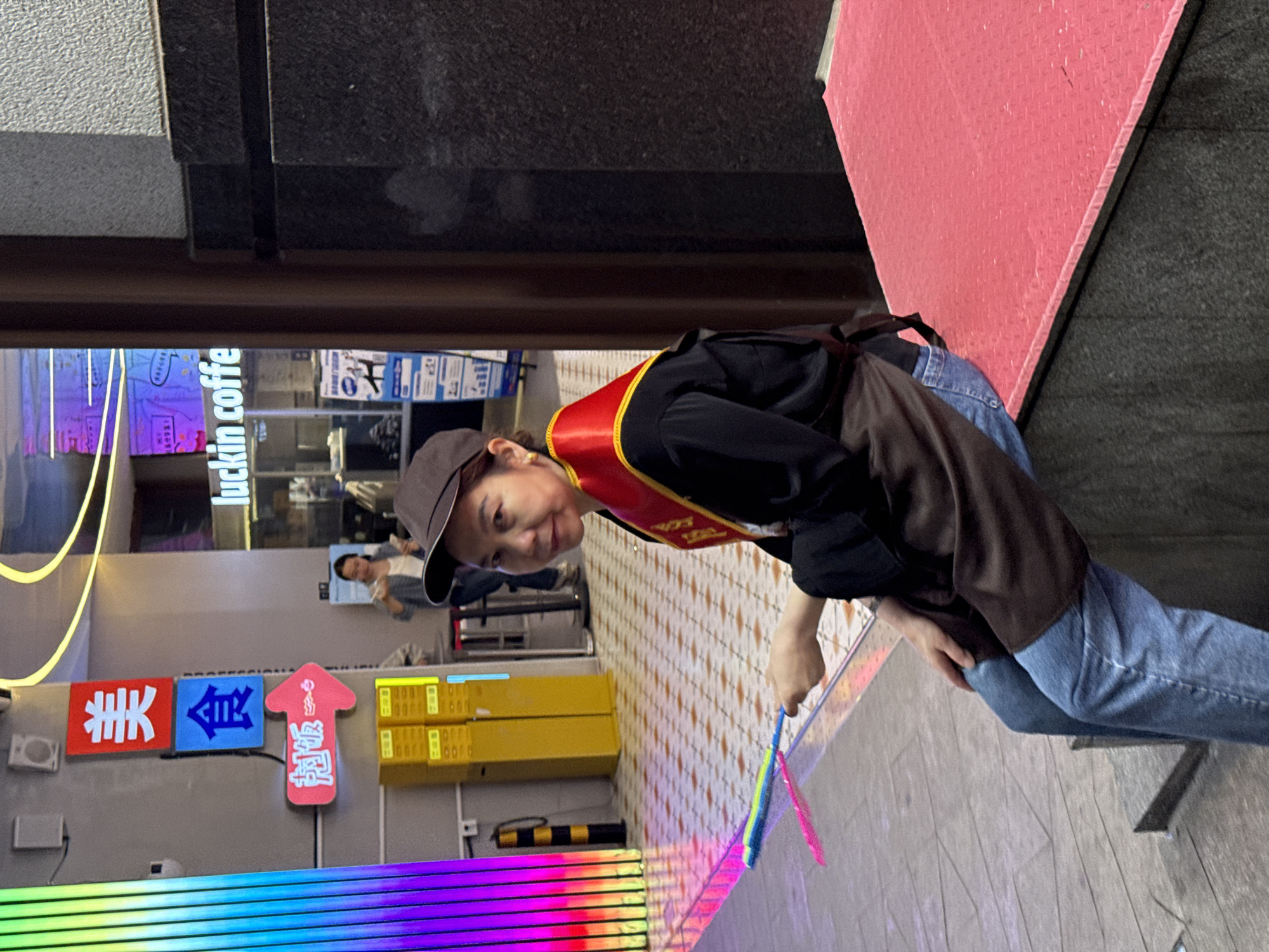 A woman wearing a brown cap, black long-sleeved shirt, brown apron, red and gold sash, and blue jeans stands on a patterned sidewalk. She is holding a colorful toy in her right hand. Behind her is a storefront with the words "luckin coffee" visible, along with signage in Chinese characters. The location appears to be in Yuzhong District, China, based on the language used on the signage and the style of architecture.