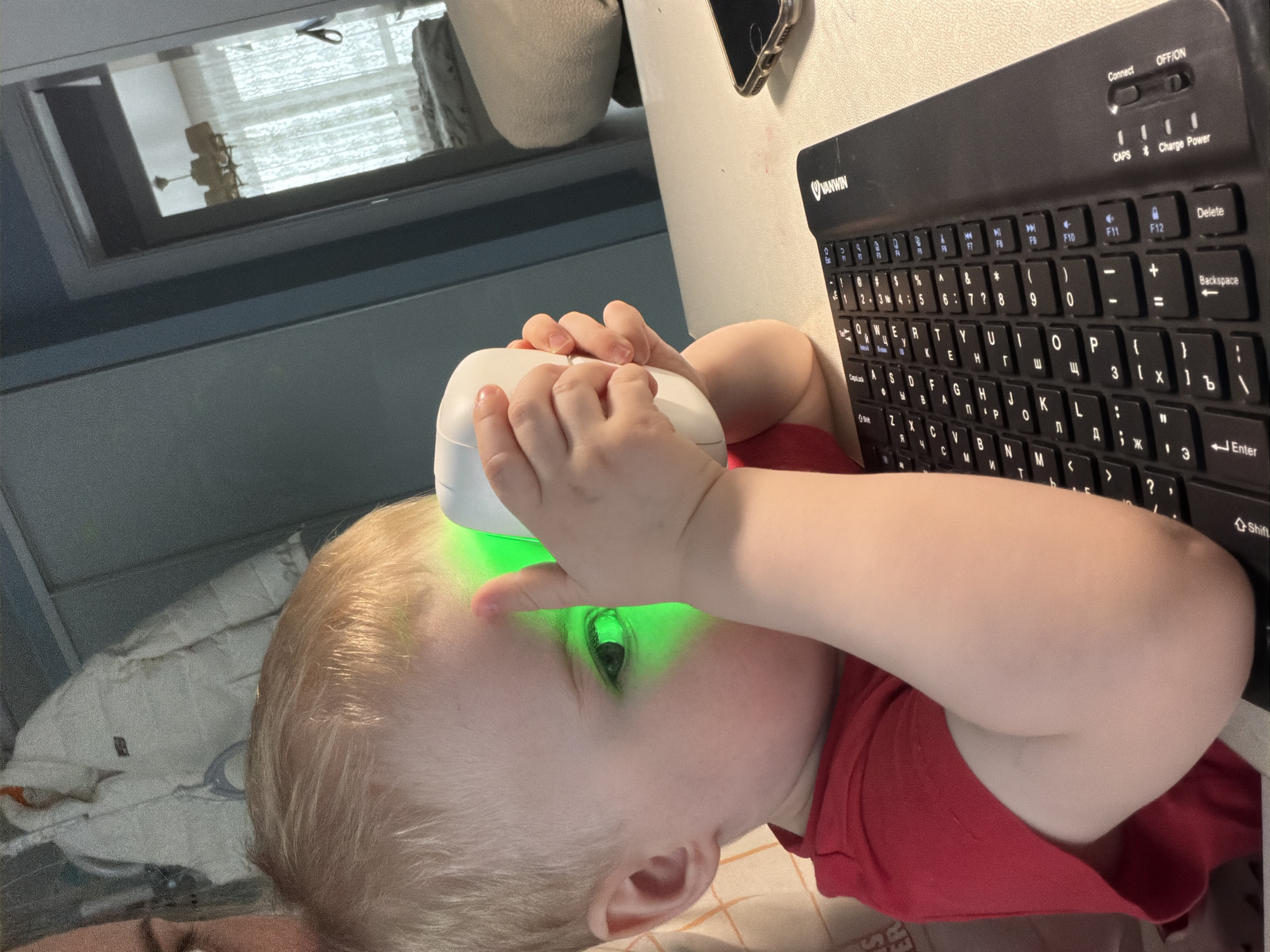 A young child with blond hair is holding a white object with a green light emitting from it close to their face. The child is wearing a red shirt. A black Vanwin keyboard with Cyrillic and Latin characters is visible next to the child. The scene appears to be indoors, with a window in the background. There is no information to ascertain that the location is Votkinsk, Russia.