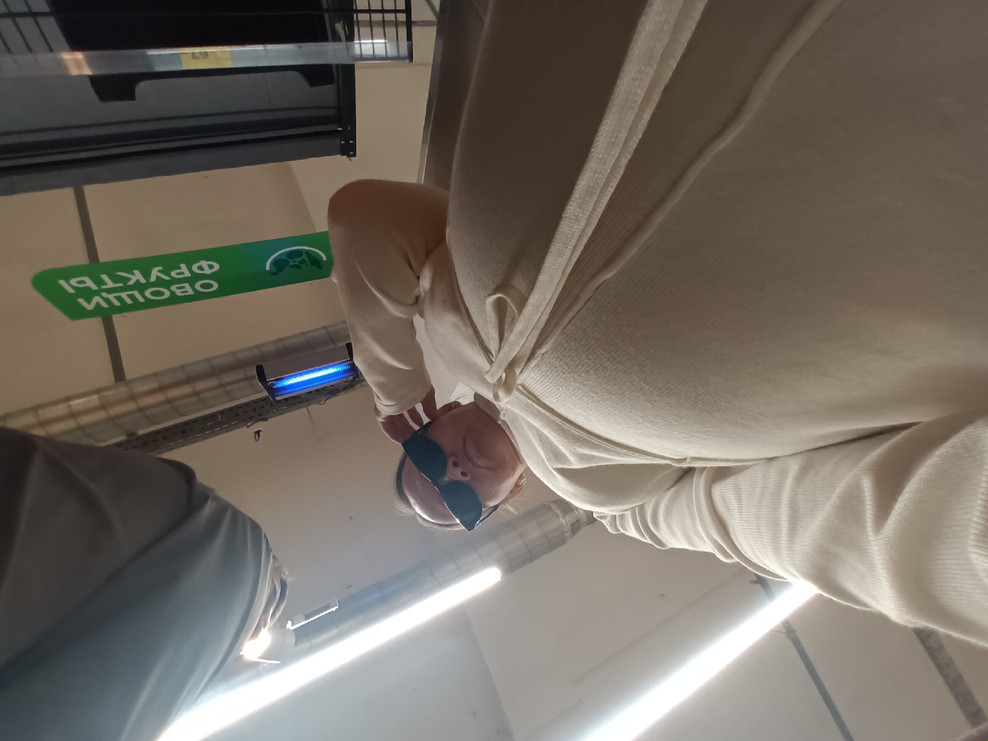 A low-angle shot depicts a woman with light skin, wearing sunglasses and a white cardigan, seemingly in a grocery store. A green sign with the Russian words "ОВОЩИ ФРУКТЫ" (Vegetables Fruits) is visible in the background. The location is likely Chaikovsky, Russia, based on the language of the sign.