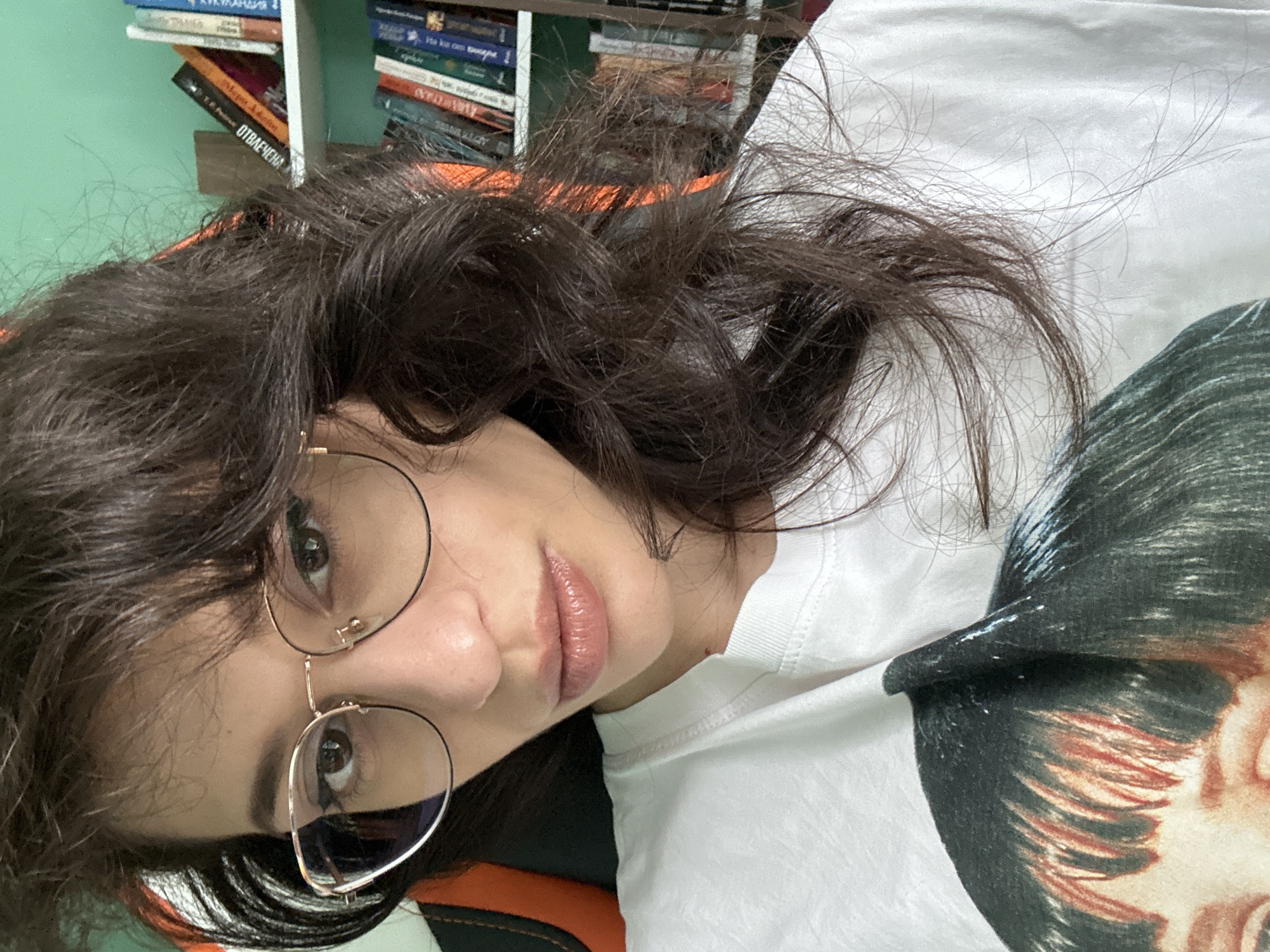 This is a vertical, close-up selfie of a young woman with dark, wavy hair and glasses. She is wearing a white t-shirt with a graphic print of a person's head and shoulders. The woman has brown eyes and appears to be looking directly at the camera. She is wearing light-colored lipstick. The image is taken indoors, with a bookshelf filled with books visible in the background. The overall mood of the image is casual and personal. The woman's hair is somewhat disheveled, and she is wearing round, thin-framed glasses. The lighting suggests it is daytime, possibly indoors with artificial light.