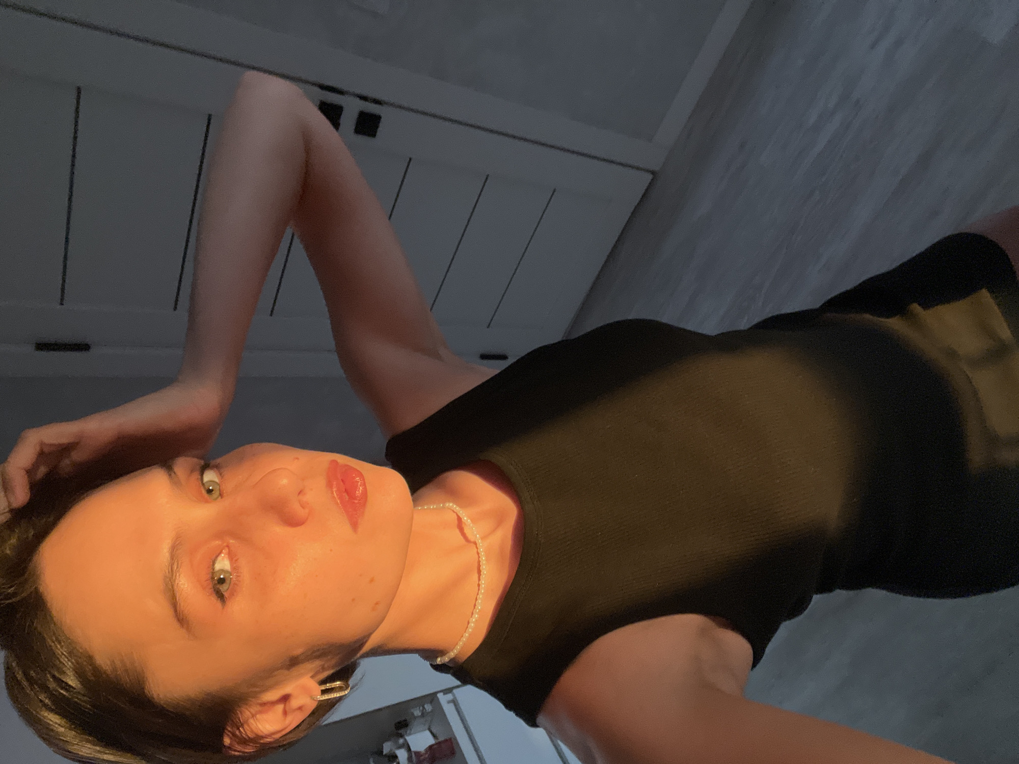 A woman is lying down, looking at the camera. She is wearing a black top and a pearl necklace. Her hair is short and dark. She has light skin and green eyes. There's a door in the background and what appears to be a floor. The location in the city of Izhevsk, Russia is unknown.