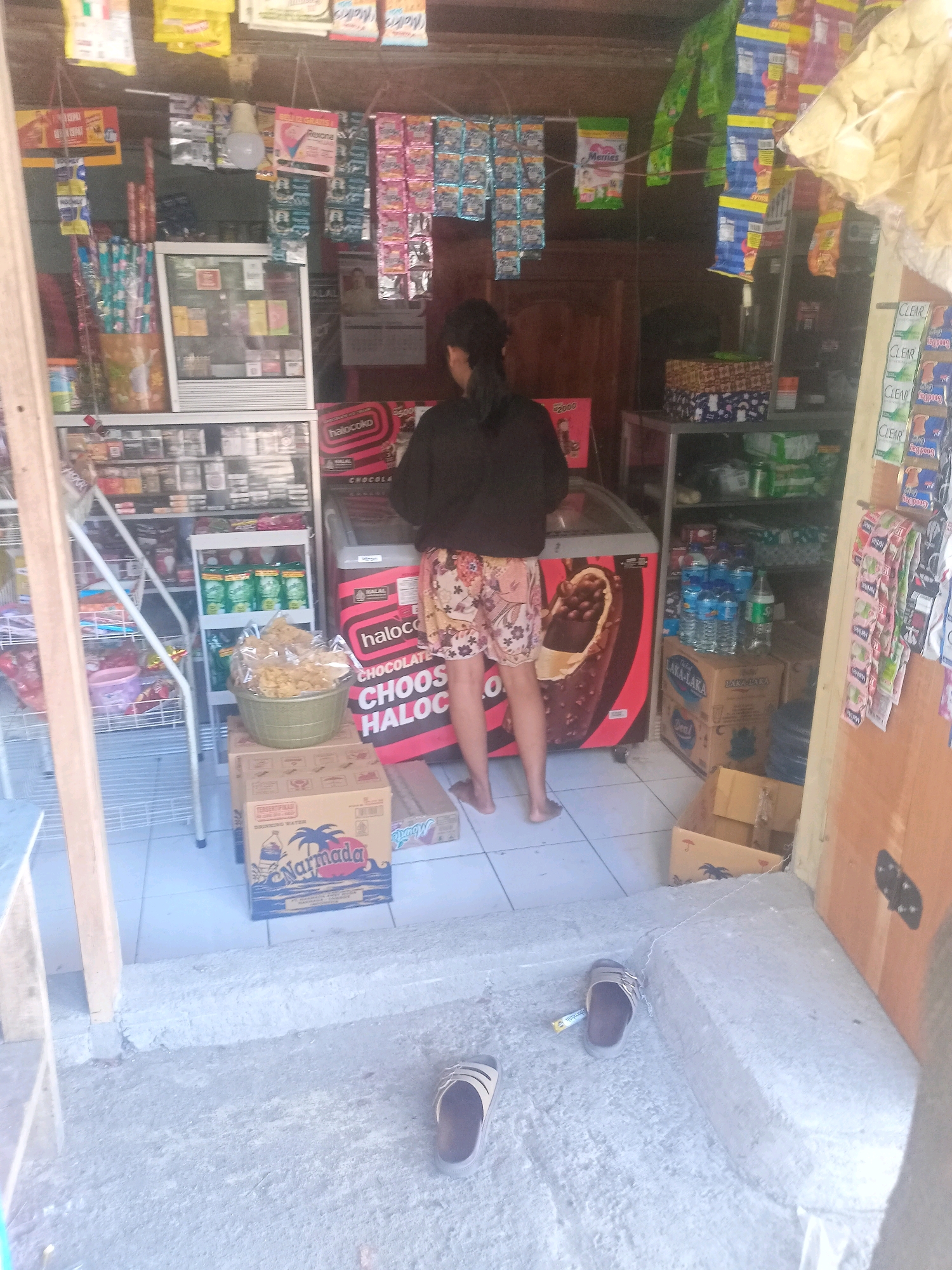 The image captures a scene inside a small convenience store, likely in Praya, Indonesia, with a focus on a display of food and beverages. A woman, partially visible from the back, is standing in front of a freezer unit which features a large image of an ice cream bar. She is wearing a dark long-sleeved top and patterned shorts. Various products are on display, including packaged snacks, drinks, and household items, arranged on shelves and hanging from the ceiling. Notably, several boxes of bottled water are stacked on the floor. Two pairs of flip-flops are visible on the ground, one near the entrance and another to the right. The store appears to be well-stocked but somewhat cluttered, with a utilitarian aesthetic. The lighting suggests an indoor setting, possibly with natural light filtering in from the entrance.