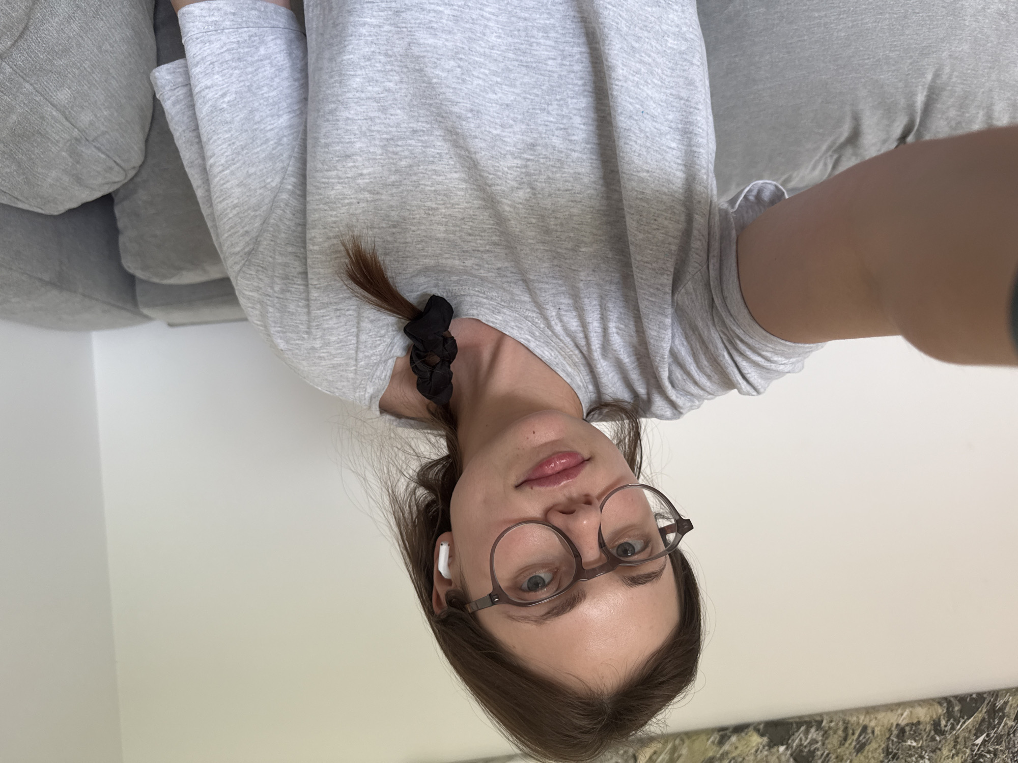 This media file is a self-portrait of a young woman with brown hair and blue eyes. She is wearing a light gray long-sleeved shirt and glasses. She has earbuds in her ears and her hair is partially tied back with a black scrunchie. The image is taken indoors, with a couch visible in the background. The lighting suggests it might be daytime. The woman appears relaxed. There is no visible text in the image.