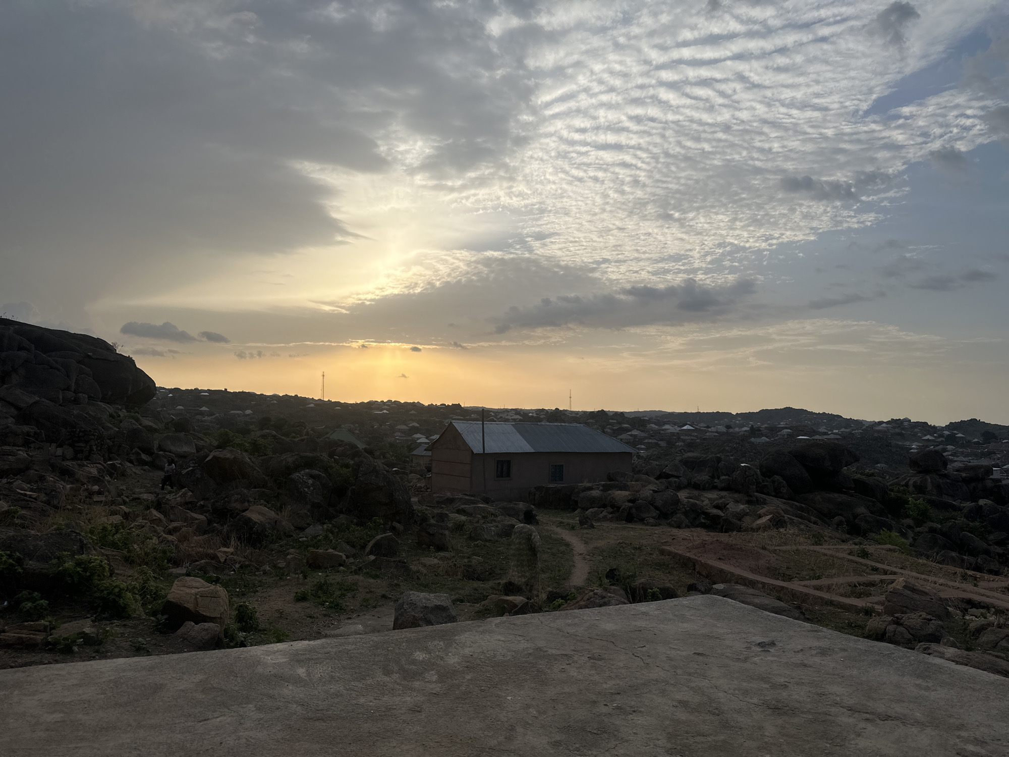 The image shows a rocky landscape with a building and a cloudy sky, likely in Jos, Nigeria. In the foreground is a concrete surface. A path leads to a building situated in the midground, surrounded by rocks and sparse vegetation. The background features a hazy sky with the sun breaking through the clouds, and the faint silhouette of a town on a distant hill. The overall scene suggests a remote or elevated location near Jos.