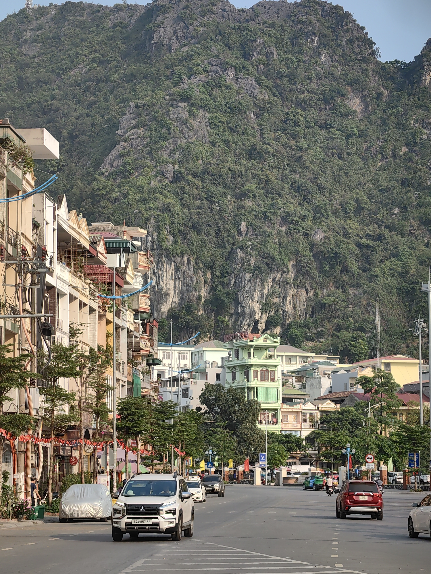 The image captures a street scene in Hong Gai Ward, Vietnam, set against a backdrop of a lush, mountainous landscape. The foreground features a multi-lane road with a white Mitsubishi SUV leading the traffic flow. A red SUV follows behind. Parked cars are visible on the sides of the road, including a vehicle covered with a protective sheet. A pedestrian is seen walking beside the road, close to some trees. Above street level, buildings line the roadside, showcasing a variety of architectural styles and colors. Festive decorations, including flags or banners, are strung along the street. In the background, a large, steep mountain covered in dense green vegetation rises prominently, dominating the scene. Some buildings are nestled at the base of the mountain. The sky appears partly cloudy or hazy.