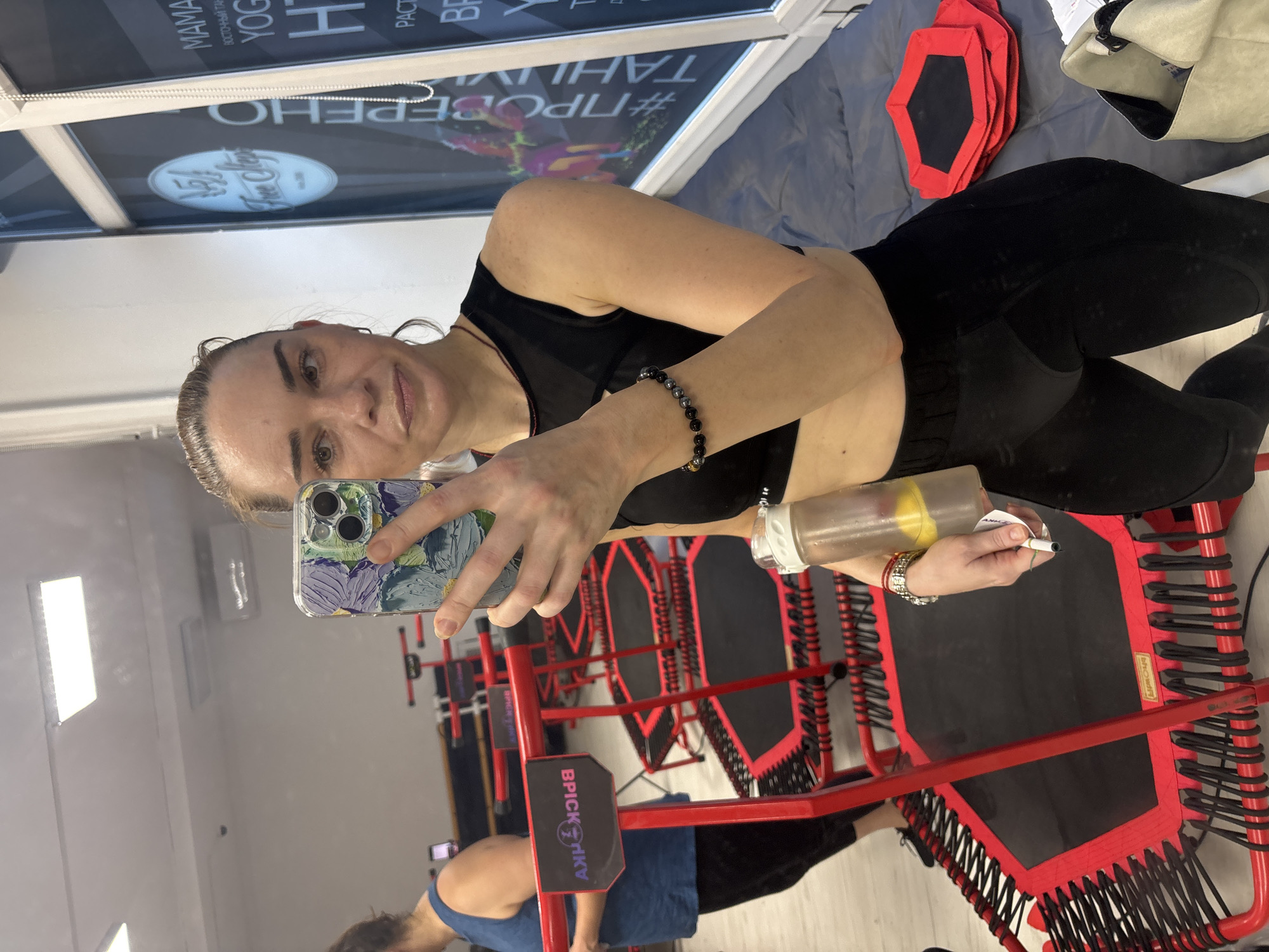 The image shows a woman in athletic wear taking a mirror selfie in a gym. She is holding a smartphone in her right hand and a water bottle in her left. The gym appears to be equipped with red mini-trampolines, some of which are visible in the background. A poster with Russian text is visible on the wall behind her. The woman is wearing a black sports bra and black leggings. She has bracelets on her left wrist. The environment suggests an indoor fitness studio.