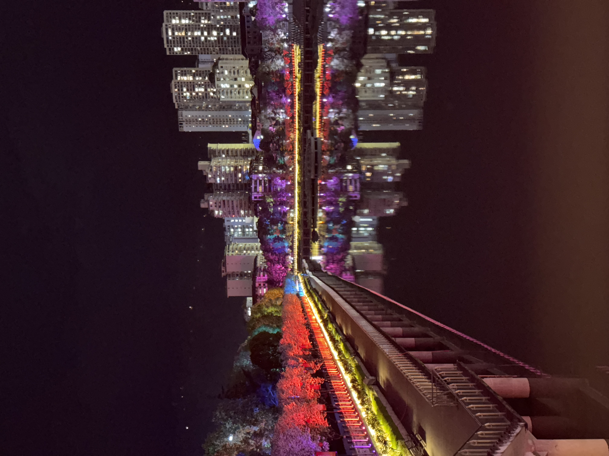The image depicts an urban landscape at night, reflected in water, likely a river or lake. The buildings in the background, adorned with lights, create a mirror effect. Along the right side of the image, a long staircase with a walkway is visible. The staircase is illuminated with a series of colored lights, ranging from red to yellow. The walkway is bordered by green vegetation. The scene evokes a sense of urban serenity and modernity in Qianjiang, China.