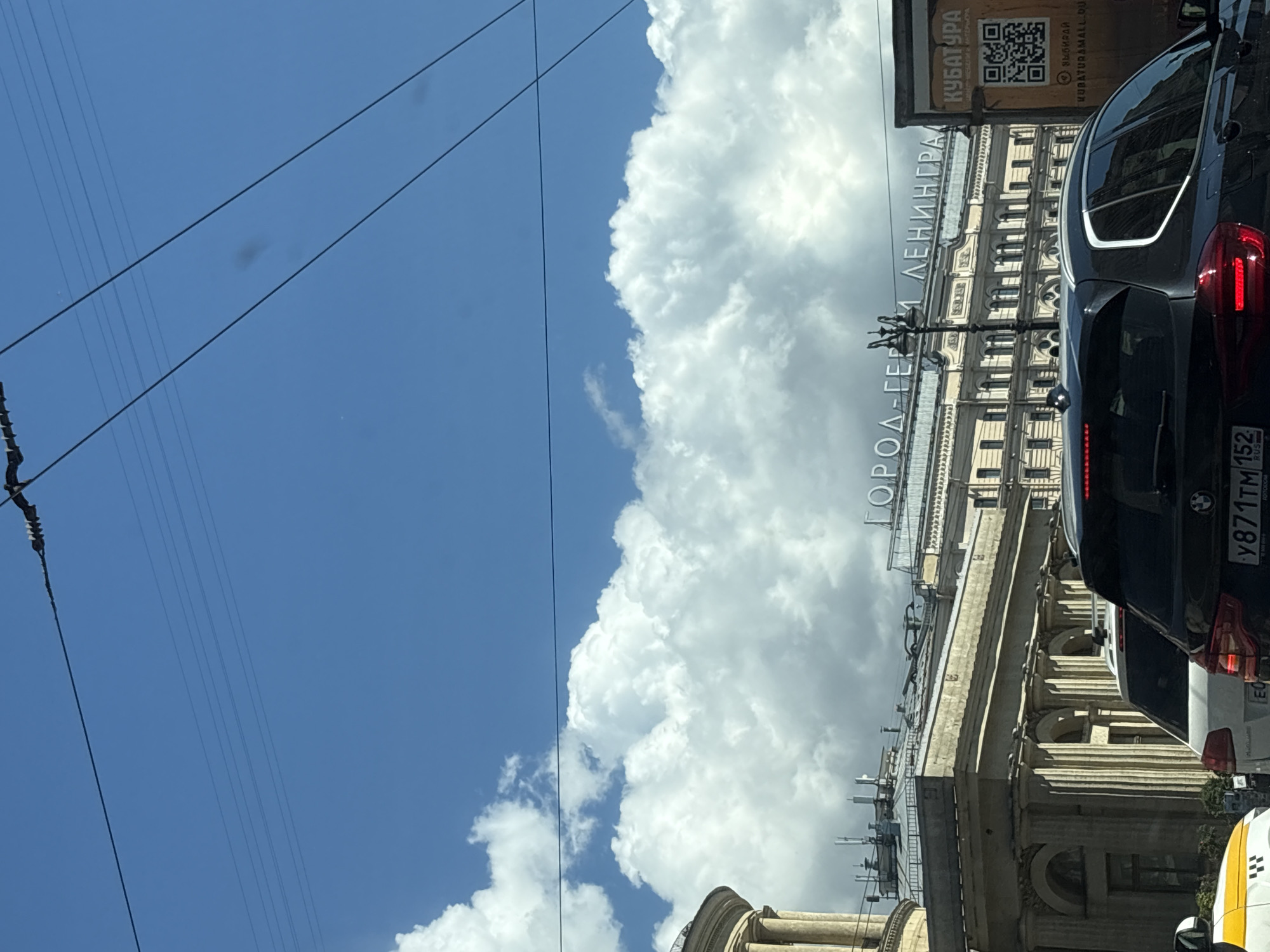 The image captures a street view in Saint Petersburg, Russia, likely on a sunny day with dramatic clouds. In the foreground, the backs of several cars are visible, including a dark SUV with illuminated brake lights and a yellow and white taxi. The license plate on the SUV reads "Y871TM 152".
Dominating the mid-ground is a grand, multi-story building with neoclassical architecture, featuring columns and arched windows. Above this building, large illuminated text spells out "ГОРОД-ГЕРОЙ ЛЕНИНГРАД" (City-Hero Leningrad), a designation historically given to cities that defended themselves bravely during World War II.
To the right of the building, a billboard displays an advertisement with a QR code and the text "КУБАТУРА" and "ВЫБИРАЙ MUBATURAMALL.RU". The sky above is a vibrant blue, with large, puffy white clouds creating a stark contrast. Overhead, power lines are strung across the frame. The overall impression is of a bustling urban environment set against a backdrop of historical significance and striking natural phenomena.