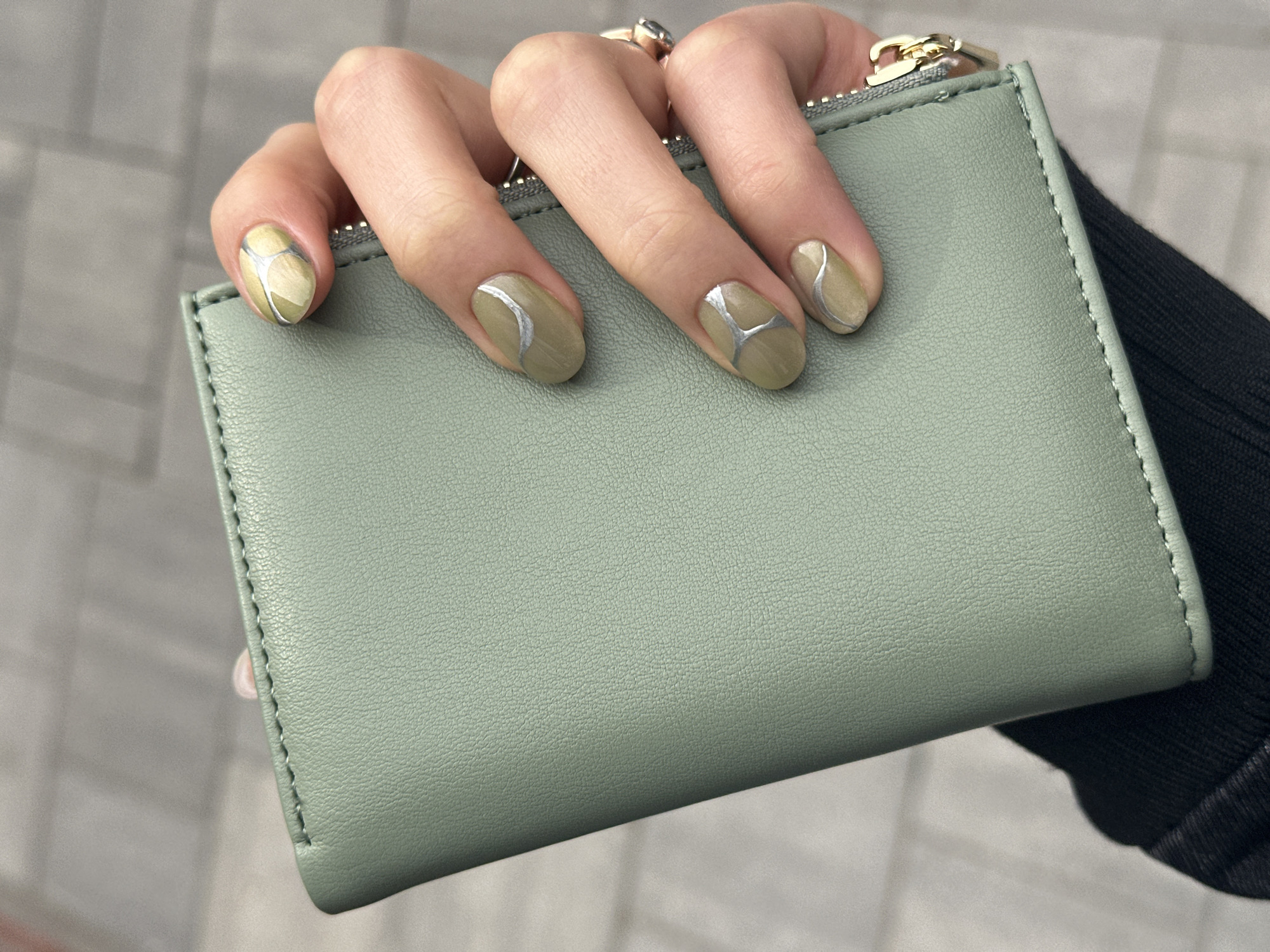 A person's hand is holding a light green leather wallet. The person is wearing rings on their fingers. The person's nails are painted with an olive green color and a silver design. The background is blurry and appears to be a light gray color, suggesting an indoor setting. The picture may have been taken in Novi Sad, Serbia, but there is no concrete evidence of the location in the image.