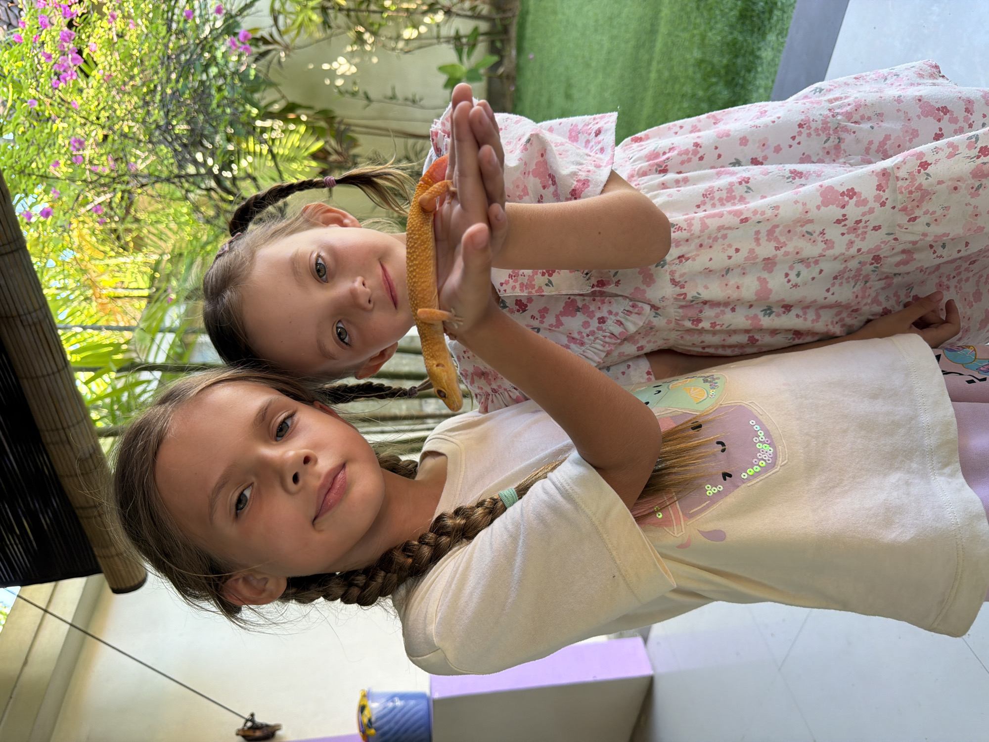 Two girls are posing with an orange lizard, likely a gecko. The girl in the foreground wears a white t-shirt with a design on it and has her hair in braids. The second girl behind her wears a light-colored dress with a pink floral pattern, also has her hair in braids, and holds out her hands, on which the gecko rests. The scene has natural light and foliage in the background, suggesting an outdoor setting. Some indoor architecture is also present with light blue surfaces. Given the location as Kuta, Indonesia, it's plausible the setting is a resort or residence with a garden area.