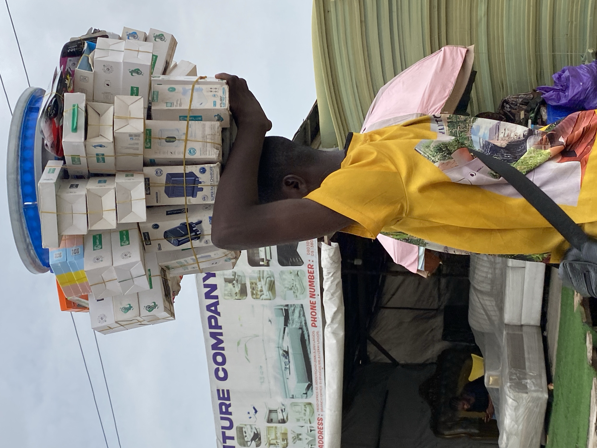 An outdoor scene in Ado, Nigeria, depicts a man balancing a large stack of small white boxes on his head. The boxes are secured with rubber bands and appear to contain phone charger adapters, labeled “Super Fast Charger.” The man is wearing a yellow shirt with a design that appears to be a collage of images. To the right of the man is a furniture store, partially visible, with a sign reading "ITURE COMPANY" and an address indicating a location in Lekki/Ajah, Lagos. Other items, including a pink object that may be a tent or umbrella, are near the furniture store display. The man is positioned so that the boxes are directly above his head, and he is holding them in place with both hands. The background shows a cloudy sky.