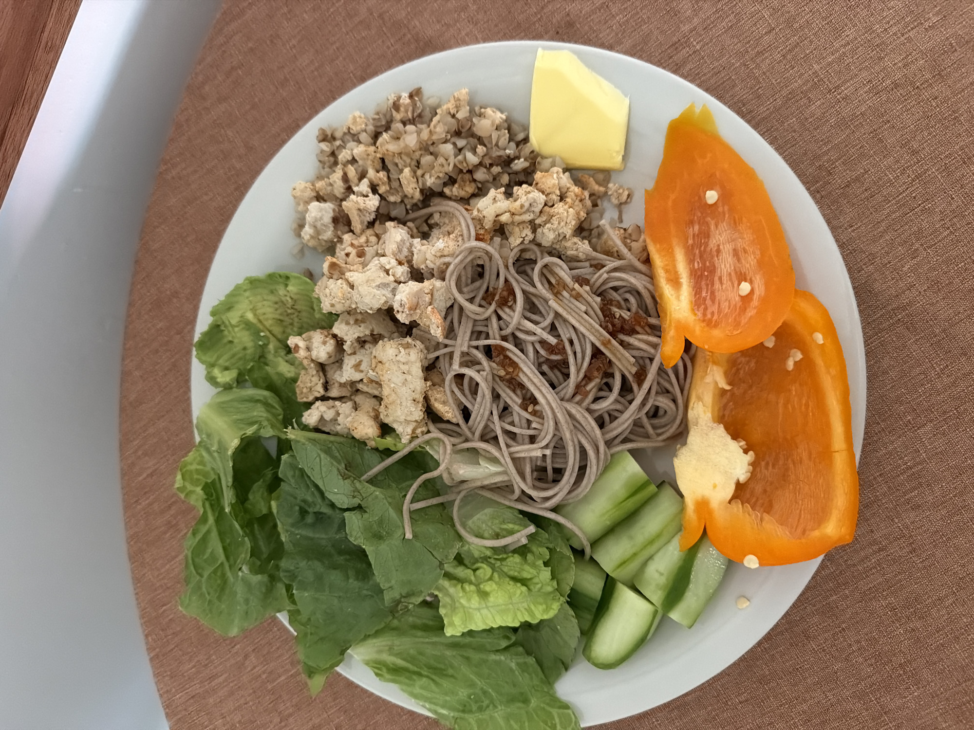 A plate of food sits on a brown surface. The plate contains buckwheat groats, a pat of butter, soba noodles, what appears to be sauteed tofu, orange bell pepper slices, cucumber spears, and lettuce leaves. A portion of a white chair can be seen on the left side of the image.
The location in Solana Beach, United States of America, cannot be ascertained solely from the content of the image. There is no contextual information to suggest where this was taken.