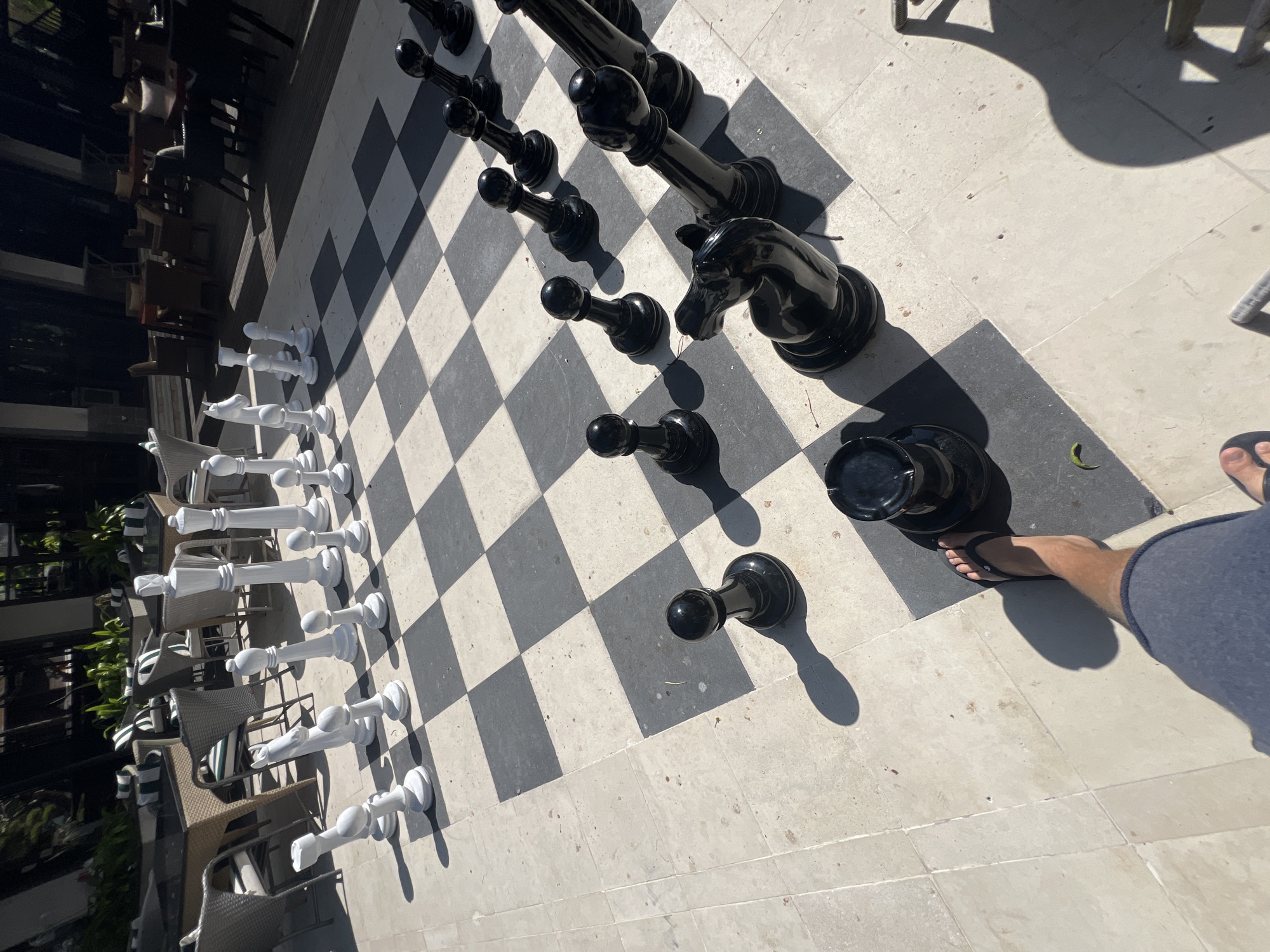 An overhead shot captures a person's legs and feet, clad in blue shorts and black flip-flops, positioned on a large outdoor chess board. The chess board has alternating black and white squares, made of stone or tile. A few of the black chess pieces, including a knight and pawns, are visible on the board, casting shadows from the bright sunlight. In the background, a shaded patio area with tables and chairs can be seen. The overall scene suggests a leisurely outdoor setting, possibly a restaurant or resort courtyard, in Denpasar, Indonesia.